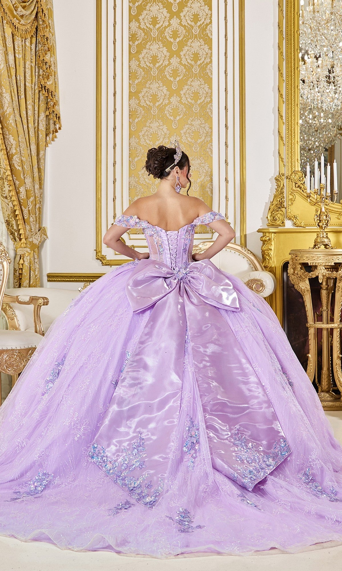 A woman in the Quinceanera Dress 5140 By Dancing Queen, a light purple gown with a large satin bow, beaded applique, and floral embroidery, stands facing away in an ornate room with gold decor.