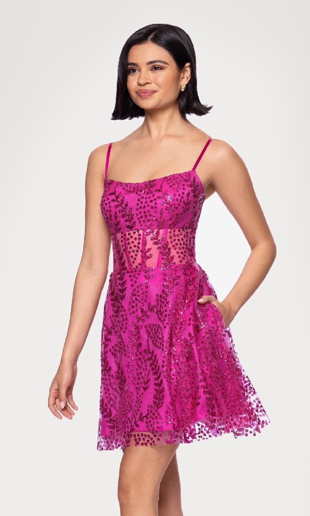 A woman models the Short Glitter Hoco Dress: Blondie Nites 5153BN—a sleeveless, bright pink cocktail dress with sheer panels and sequin accents—against a plain light background.