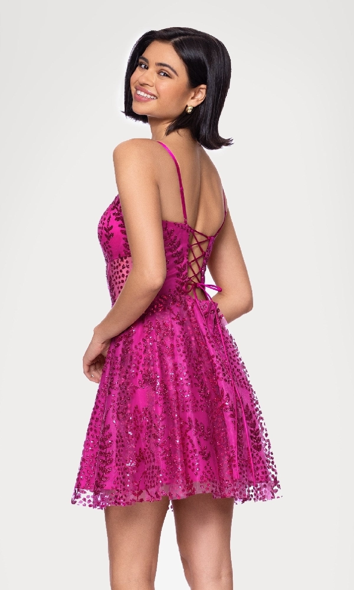 A woman smiles, turned slightly to the side, wearing the Blondie Nites 5153BN—a short glitter hoco dress in pink with sequins and a sleeveless lace-up back design.