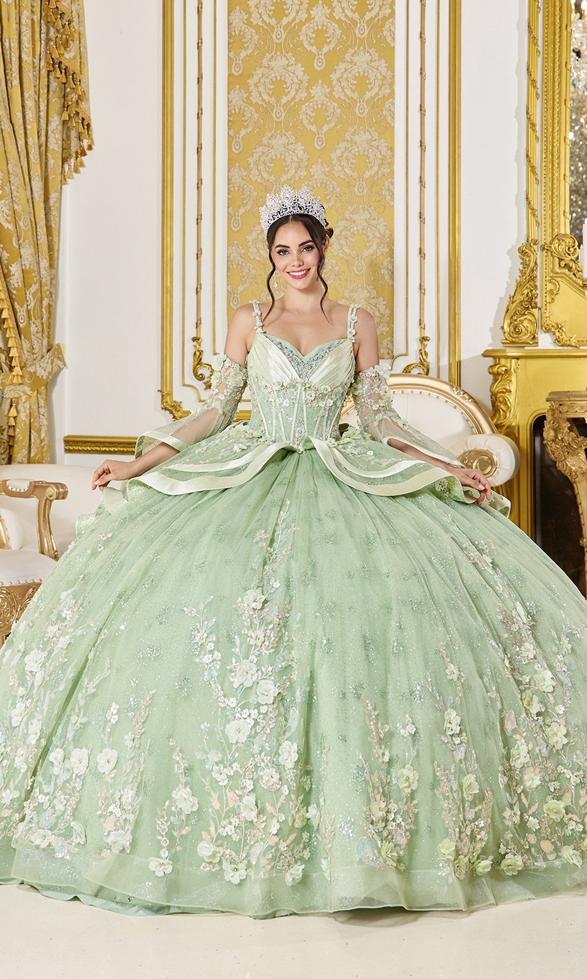 Wearing the Quinceanera Dress 5154 By Dancing Queen in elegant sage, adorned with intricate floral embellishments and a sparkling tiara, a woman sits gracefully on a white and gold ornate sofa in an opulent, gold-accented room—perfectly embodying the regal essence of this Dancing Queen Quinceanera gown.