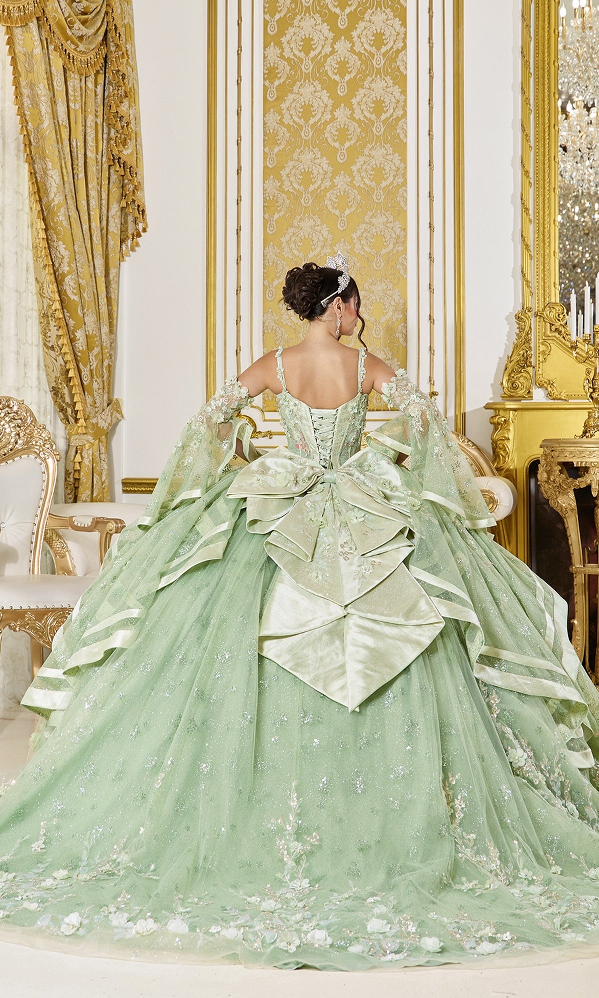 A woman stands facing away in an opulent, gold-decorated room, wearing the Quinceanera Dress 5154 by Dancing Queen—a large, ornate sage green ball gown adorned with a bow.