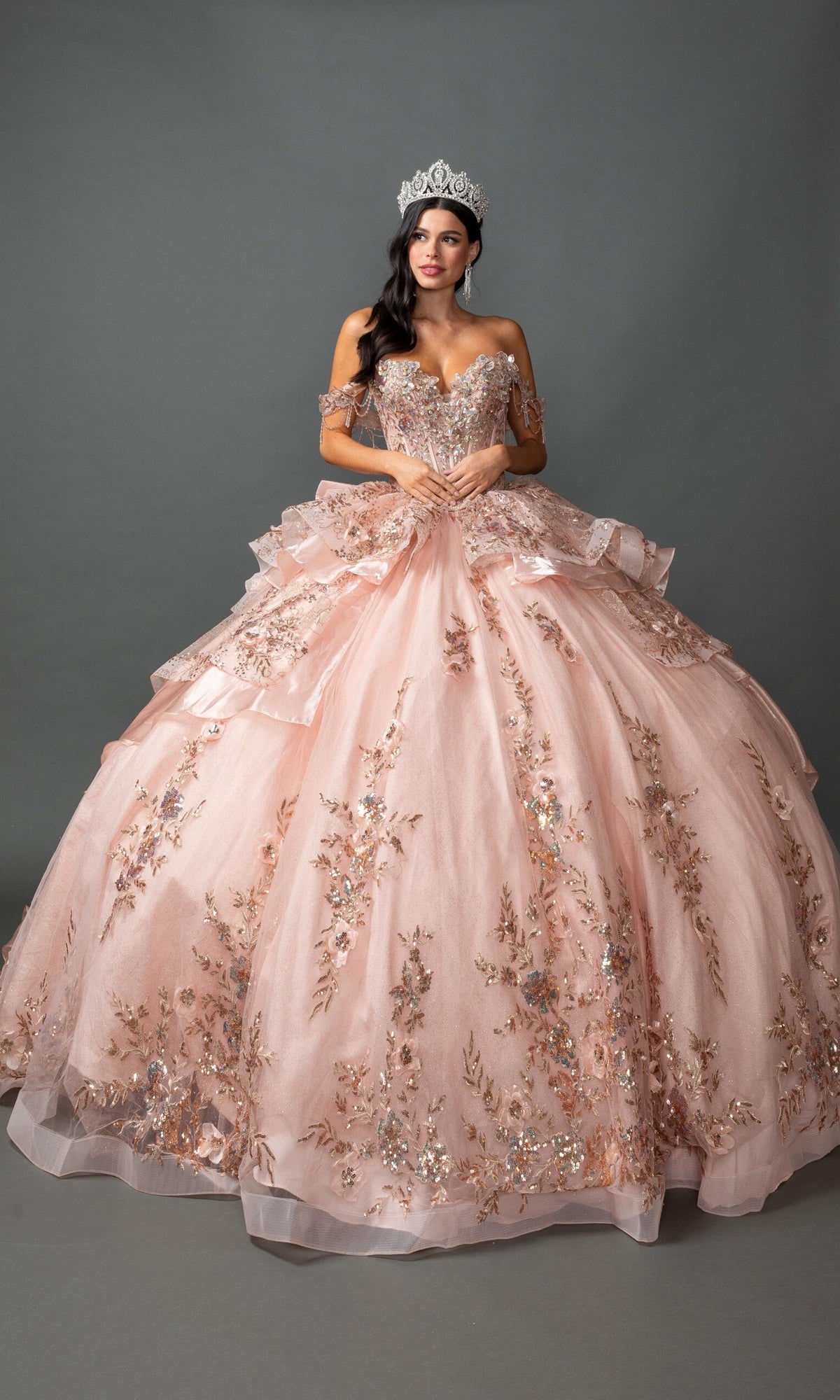 A woman stands facing forward with hands clasped, adorned with a sparkling tiara, wearing the Quinceanera Dress 5157 By Dancing Queen in rose goldโa ball gown featuring 3D floral appliques and an off-the-shoulder sweetheart necklineโagainst a plain gray background.