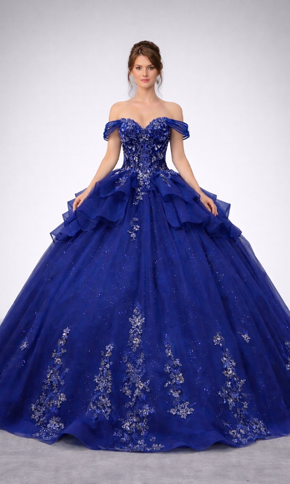 A woman wearing the Quinceanera Dress 5165 By Dancing Queen in navy, featuring an off-shoulder corset bodice adorned with floral embellishments and a full, layered skirt reminiscent of a glitter-tulle ballgown, stands against a plain background.