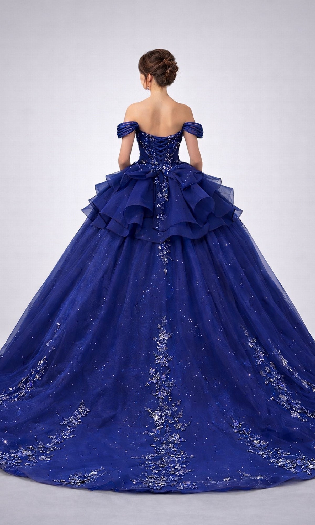 A woman with an elegant updo hairstyle wears the Quinceanera Dress 5165 By Dancing Queen, a stunning off-the-shoulder, floor-length navy-blue gown embellished with glitter and delicate floral lace appliques. She is facing away from the camera.
