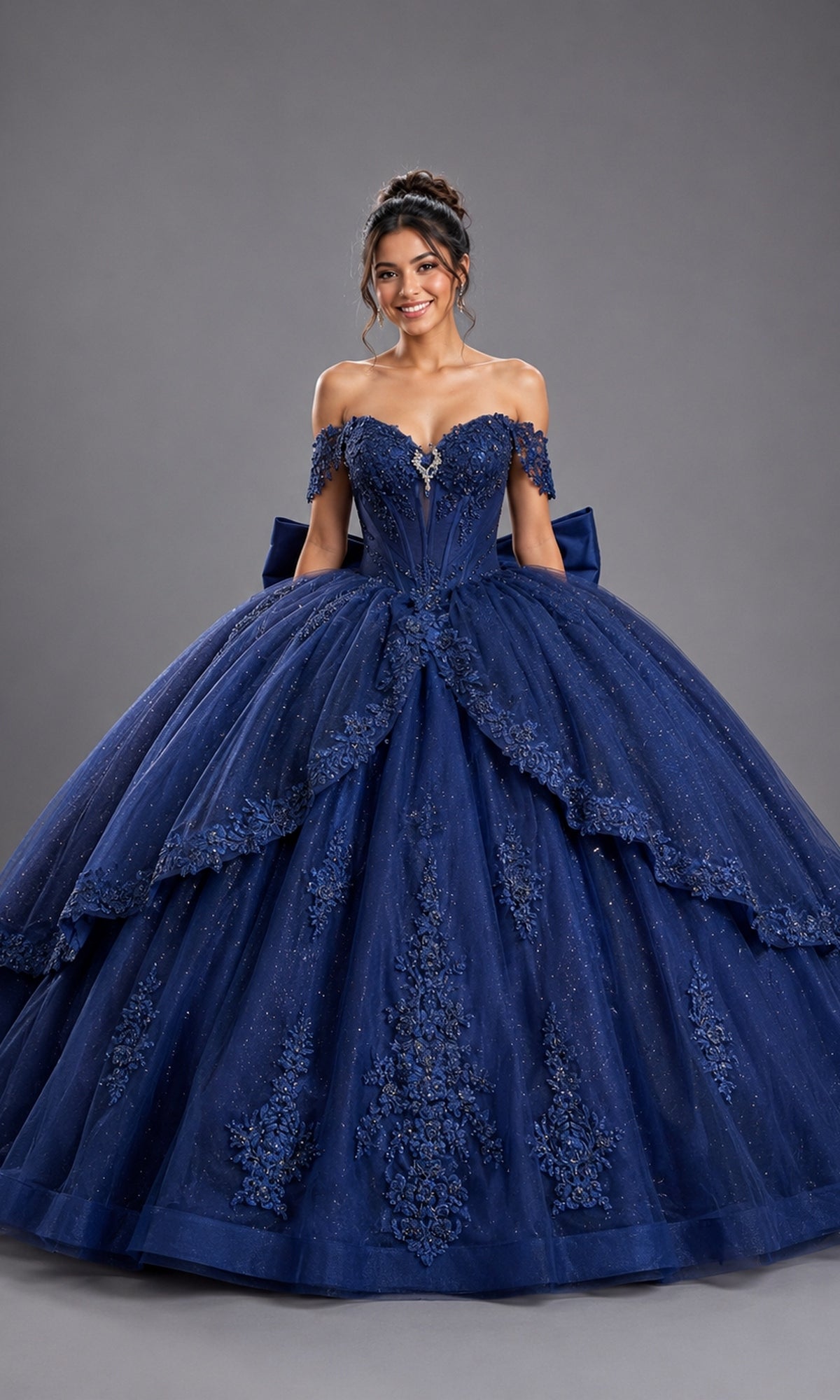 A woman stands smiling in the Quinceanera Dress 5172 by Dancing Queen—a navy, off-shoulder ball gown adorned with lace and sequin details and featuring a corset-style bodice—against a plain gray backdrop.