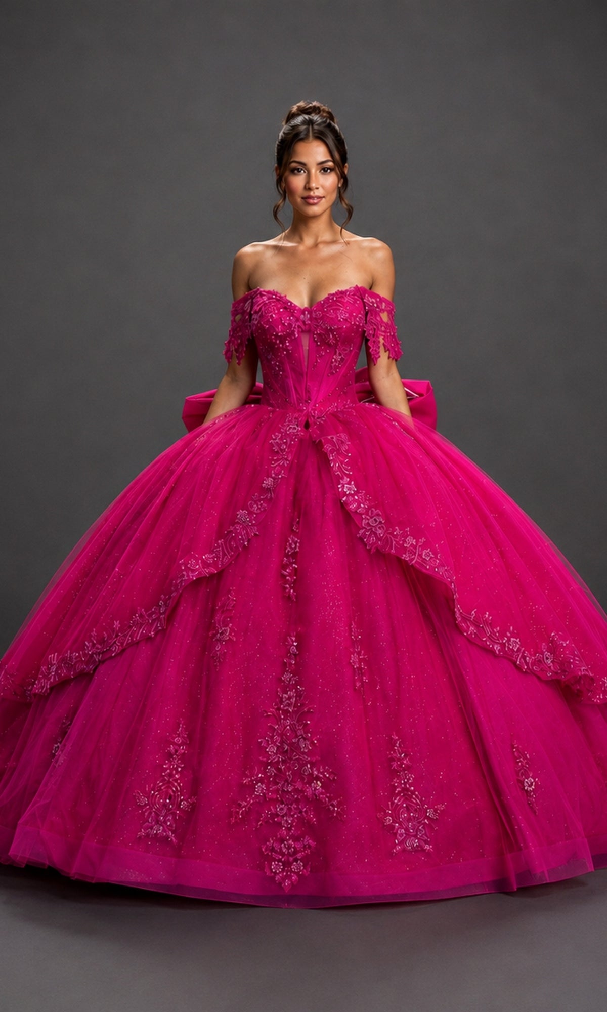 A woman wearing the Quinceanera Dress 5172 By Dancing Queen in fuchsia, featuring an off-the-shoulder corset-style bodice and a layered tulle skirt embellished with floral details, stands against a plain dark background.
