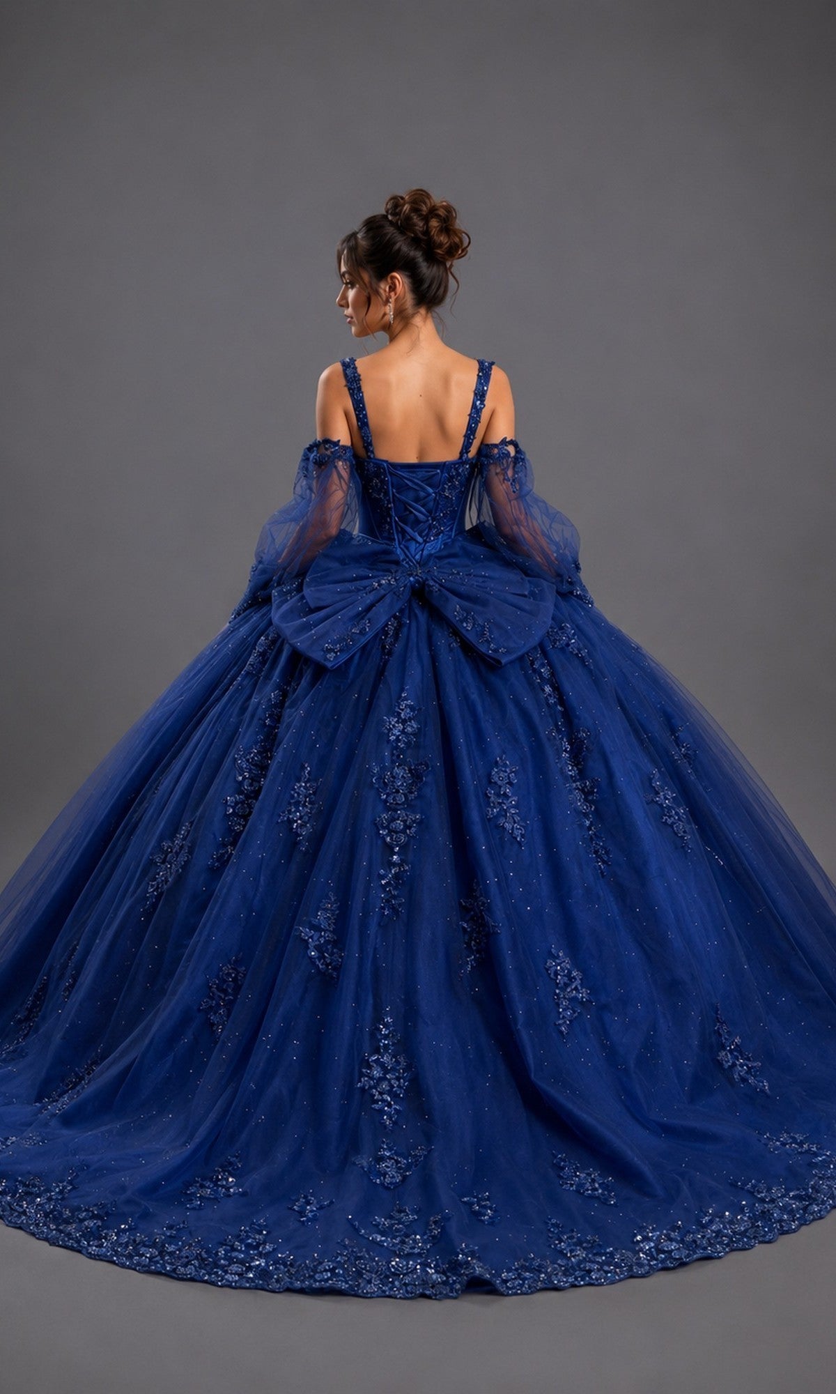 A woman wearing the Quinceanera Dress 5177 by Dancing Queen, featuring a royal blue ball gown silhouette with floral embellishments, a shimmering ballgown skirt, tulle sleeves, and a large bow on the back, stands against a plain gray background.
