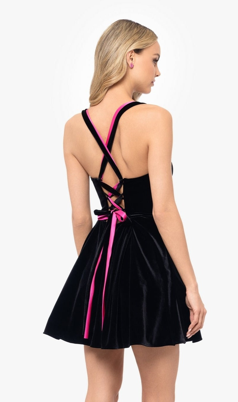 A woman stands facing away in the Pink-Ribbon Short Black Velvet Hoco Dress 5177BN, featuring a flared skirt and crisscross pink and black ribbon back detailing.