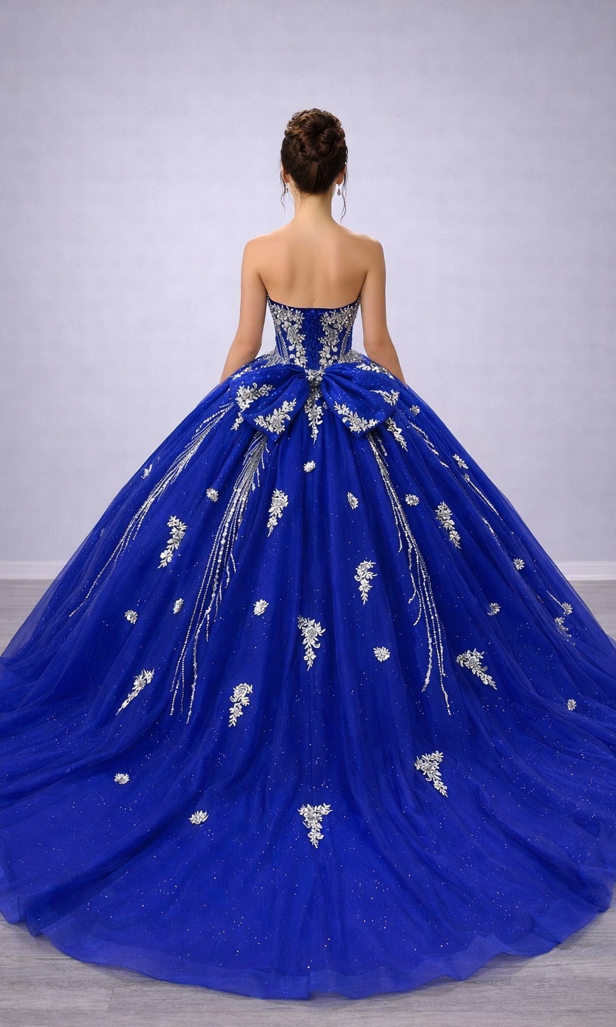 A woman stands facing away against a plain background, wearing the Quinceanera Dress 5183 by Dancing Queen—a blue strapless gown featuring silver floral embellishments and a structured corset bodice.