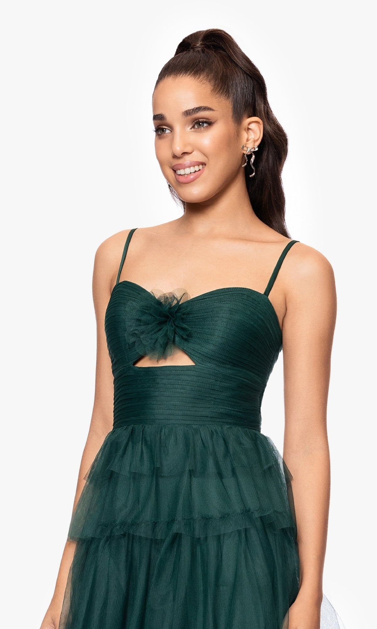 A woman stands smiling against a white background wearing the Blondie Nites 5183BN Dark Green Ruffled Prom Gown with spaghetti straps, a tulle skirt, floral accent, and a cutout bodice.