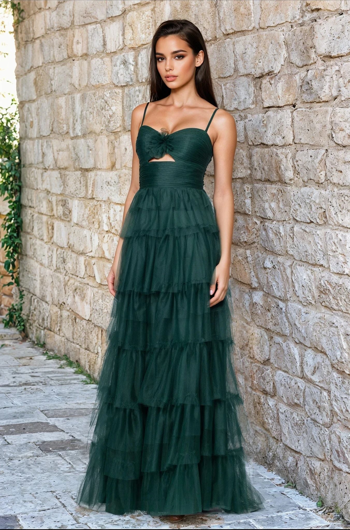Woman standing by a stone wall in the Dark Green Ruffled Prom Gown (Blondie Nites 5183BN), featuring a tiered A-line silhouette, spaghetti straps, and a front cutout.