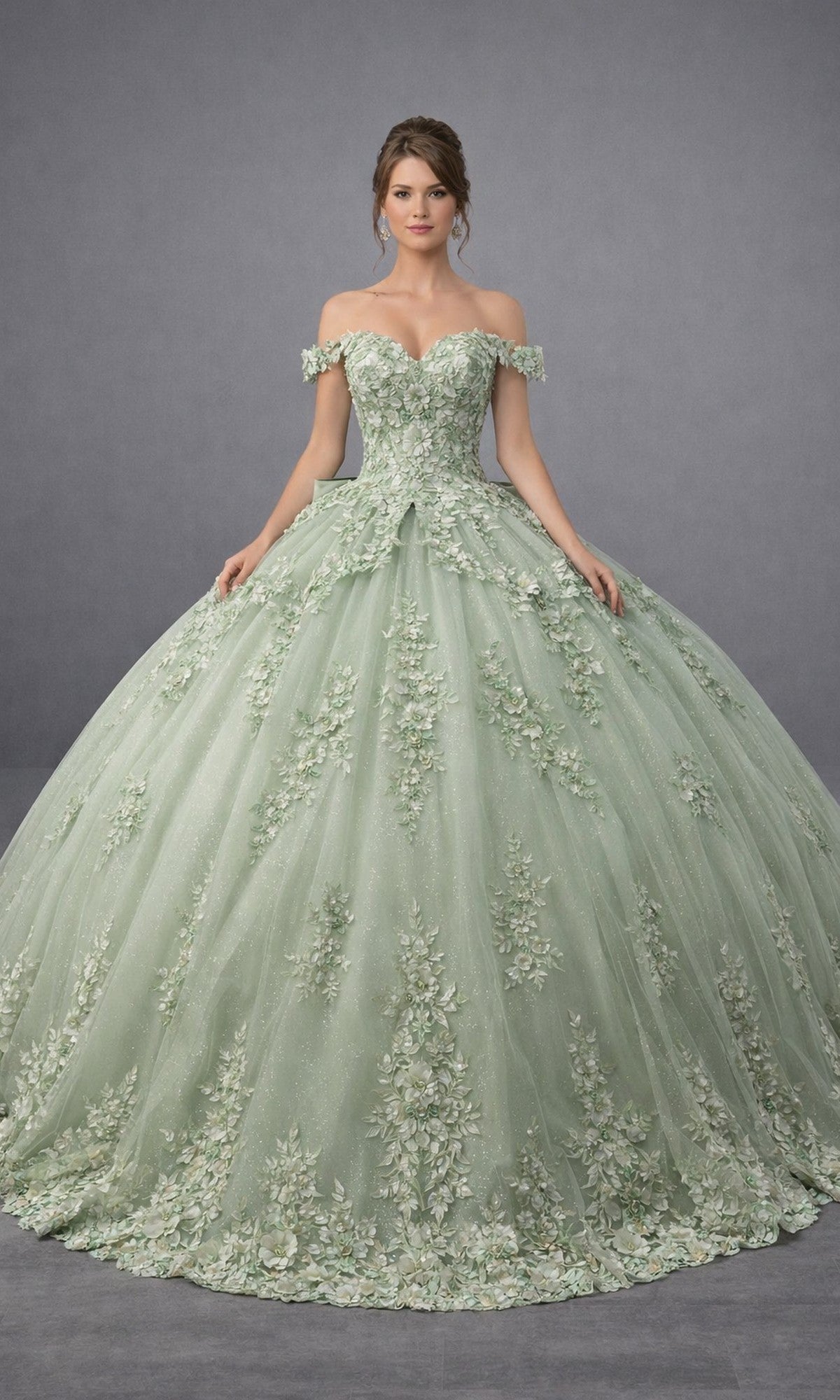 A woman wearing the Quinceanera Dress 5184 By Dancing Queen in sage, featuring an off-shoulder neckline, delicate floral embroidery, and stunning floral appliques on a voluminous ballgown skirt, stands against a plain gray background.