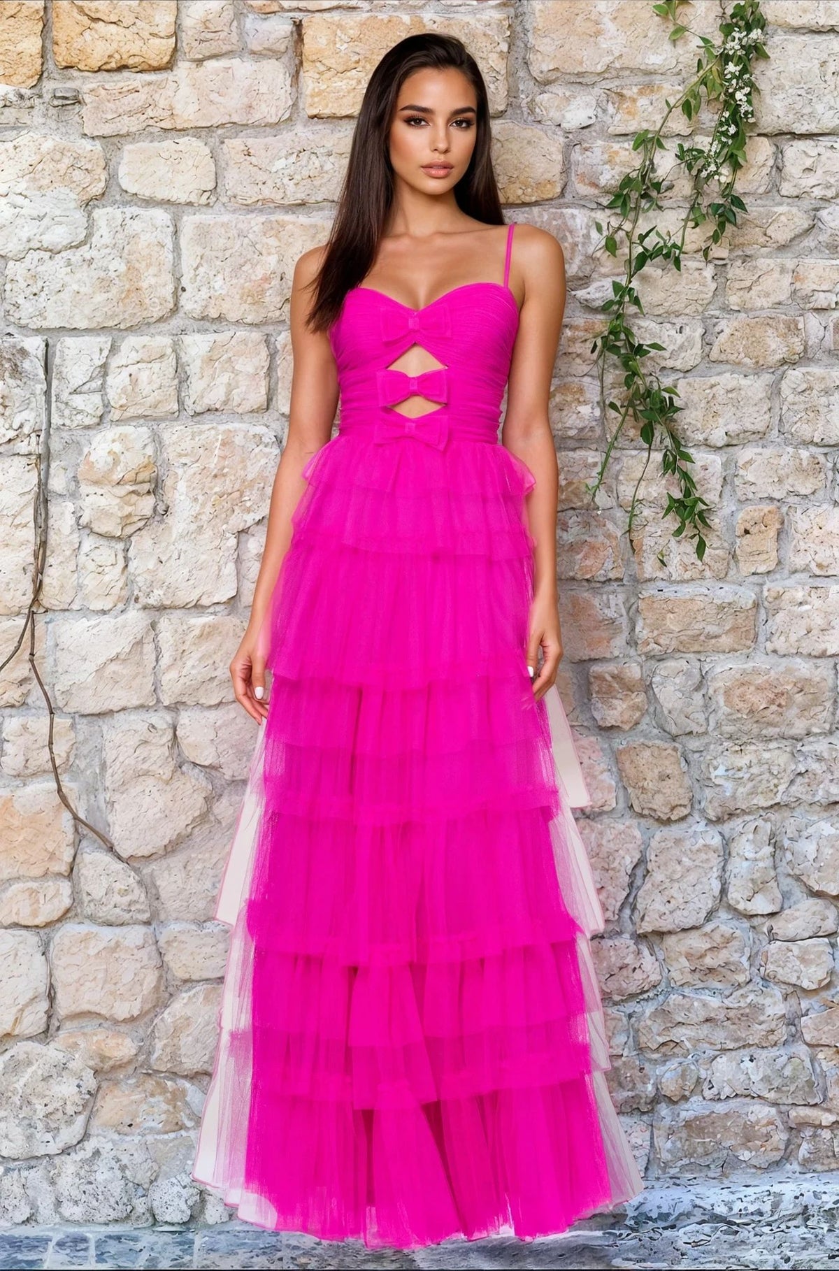 A woman stands by a stone wall in the Blondie Nites 5187BN long hot pink prom dress, featuring thin straps, geometric cut-outs, and a ruched sweetheart bodice—a standout option under $200.