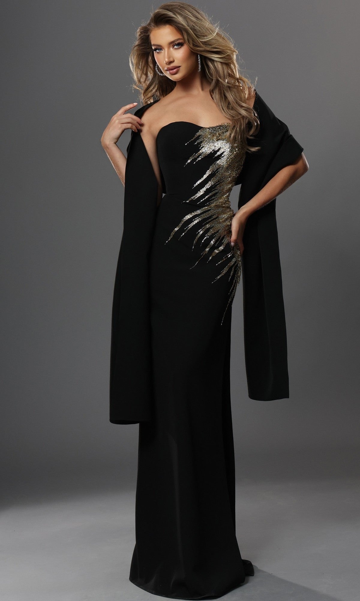 A woman is wearing the Jovani 51900 long formal dress—a black strapless gown featuring intricate gold sequin embroidery and paired with a matching black shawl—while standing against a plain gray background. The fitted sheath silhouette of the dress elegantly flatters her figure, and the sparkling gold embellishments lend an air of sophistication to her look.