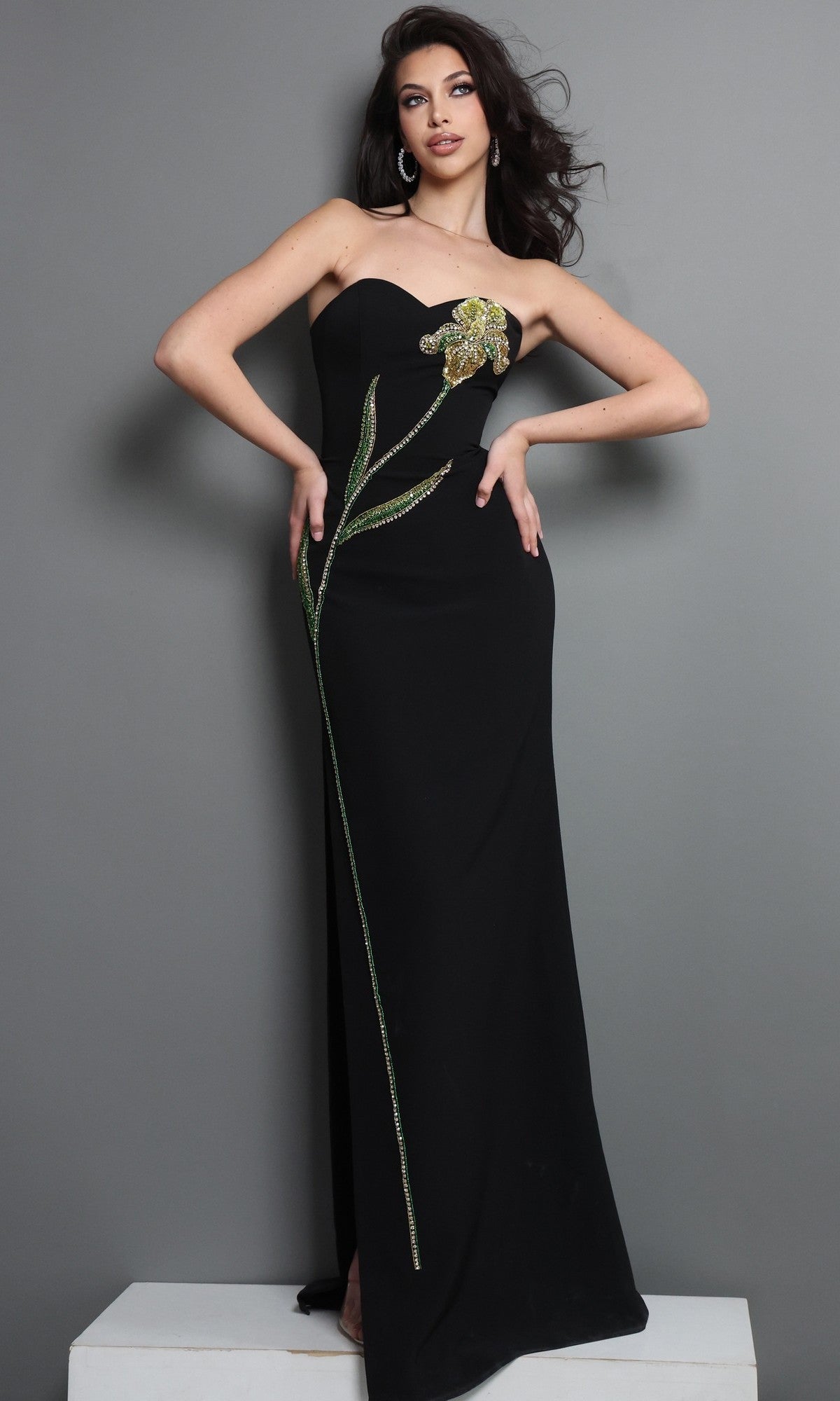 A woman wears the Long Formal Dress: Jovani 51902, a striking black strapless gown featuring intricate green and gold beaded floral embellishments, as she stands against a gray background.