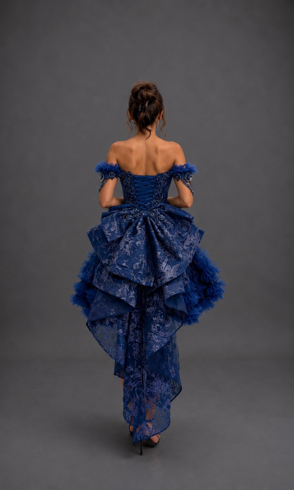 A woman stands with her back to the camera, wearing the Quinceanera Dress 5192 by Dancing Queen—a stunning blue ball gown featuring an off-shoulder layered design with intricate lace and tulle details, complemented by a detachable layered skirt, set against a plain gray background.