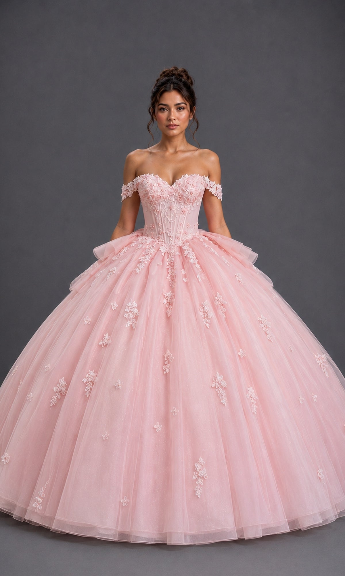 A woman wears the Quinceanera Dress 5200 By Dancing Queen in blush, showcasing delicate floral appliqué details and an off-the-shoulder sweetheart neckline as she stands gracefully against a plain gray background.