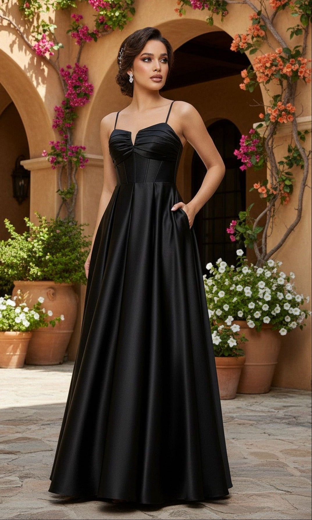 A woman stands in a courtyard with potted plants and vines, wearing the Blondie Nites 5207BN—a long black satin prom dress featuring an a-line silhouette, thin straps, and hidden pockets.