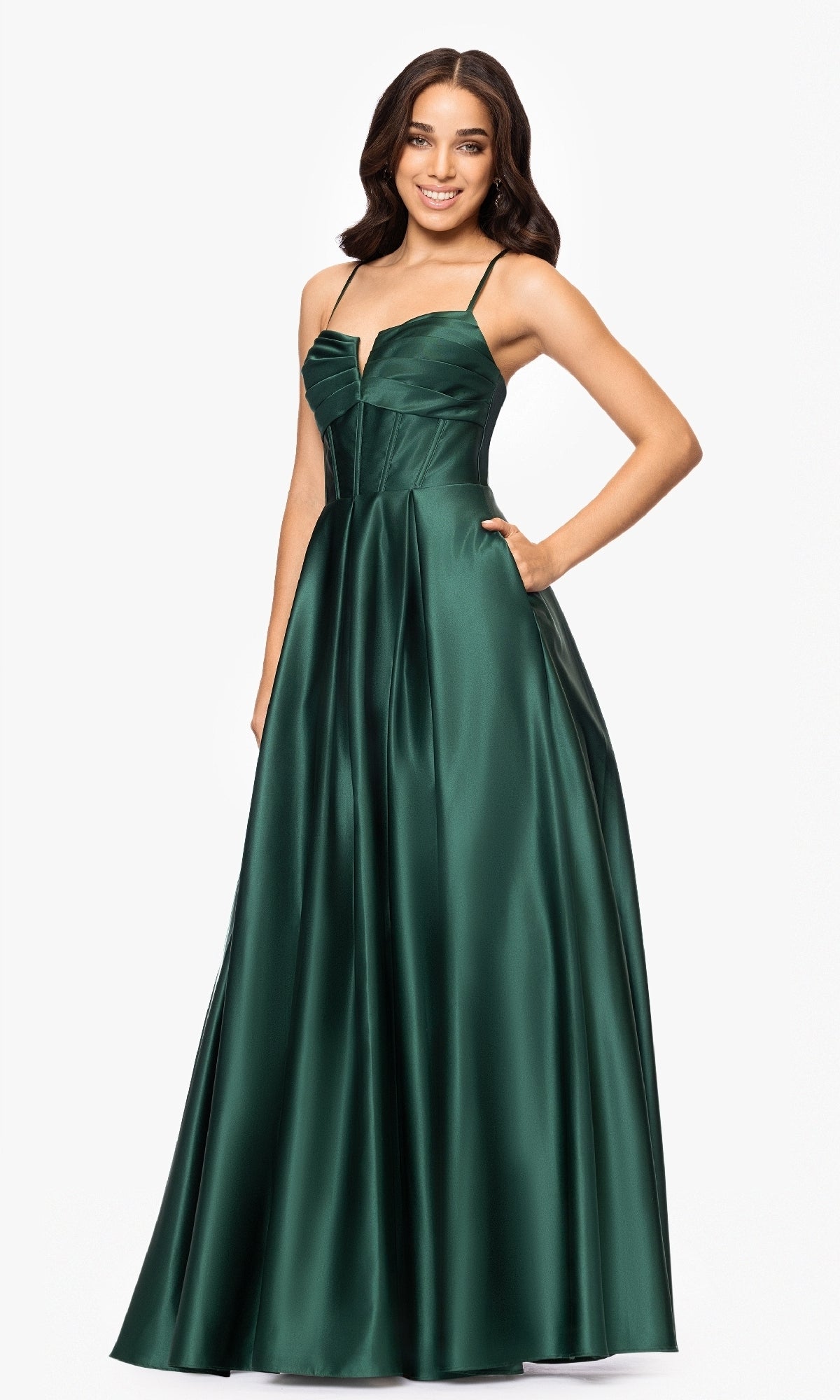 A woman in the Blondie Nites 5207BN long dark green satin prom dress, under $200, with spaghetti straps, a fitted corset bodice, a-line skirt with pockets, stands against a plain white background.