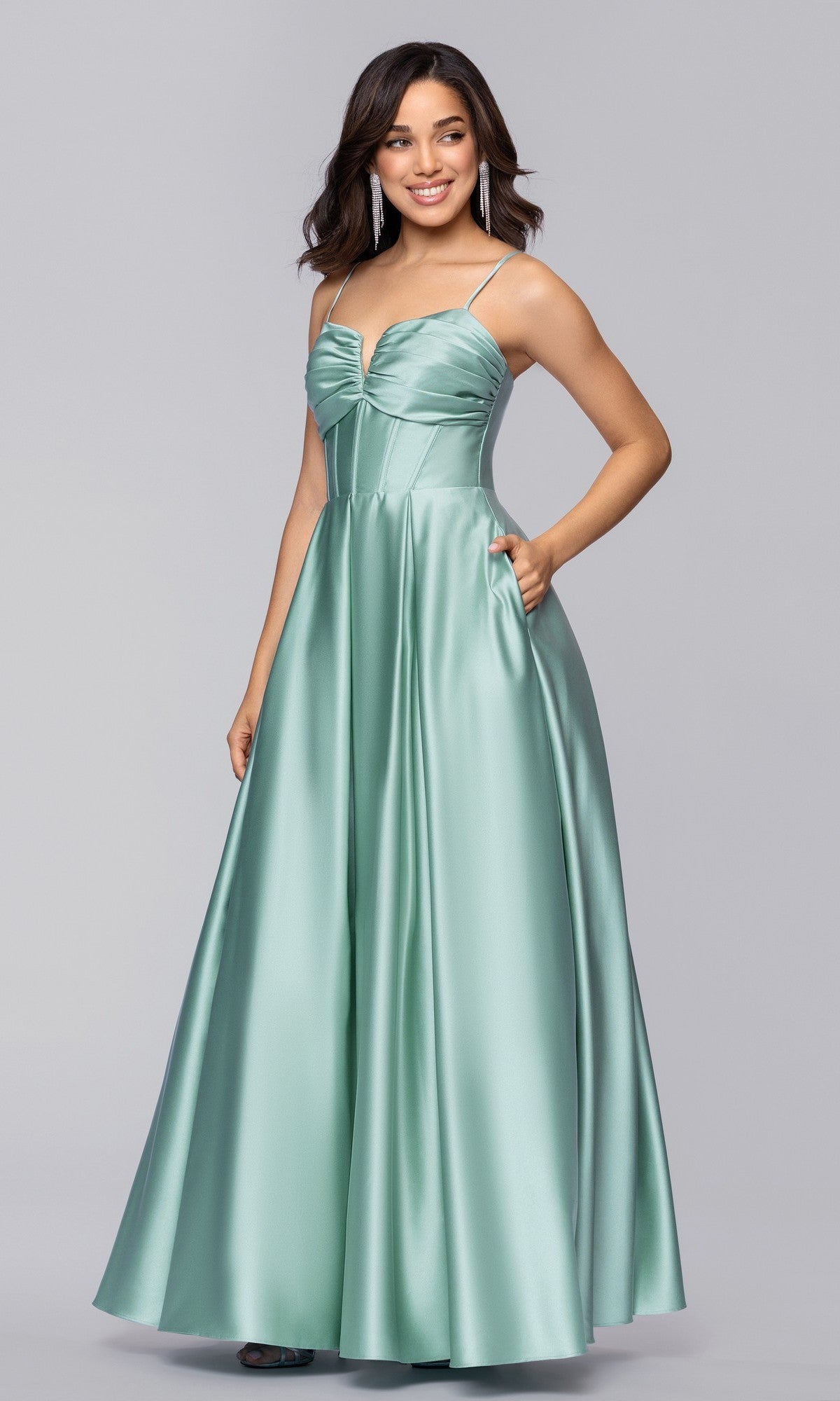 A woman wears the Blondie Nites 5207BN, a long satin A-line prom dress with spaghetti straps, ruched corset bodice, and pockets, against a plain gray background. This elegant gown is available for under $200.