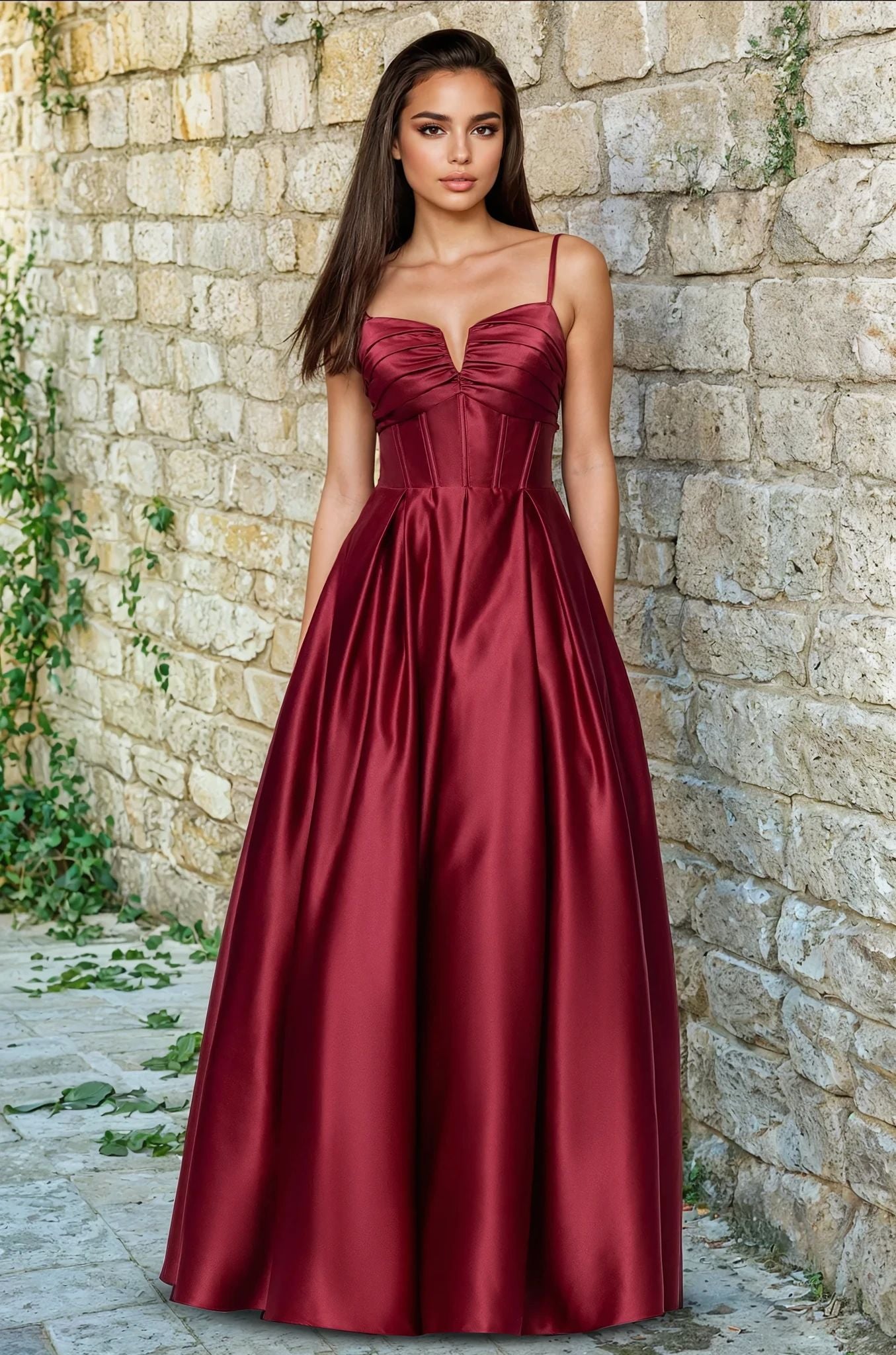 A woman stands before a stone wall in the Blondie Nites 5207BN long prom dress, featuring an elegant a-line satin silhouette, fitted corset bodice with thin straps, and a floor-length burgundy skirt—all for under $200.