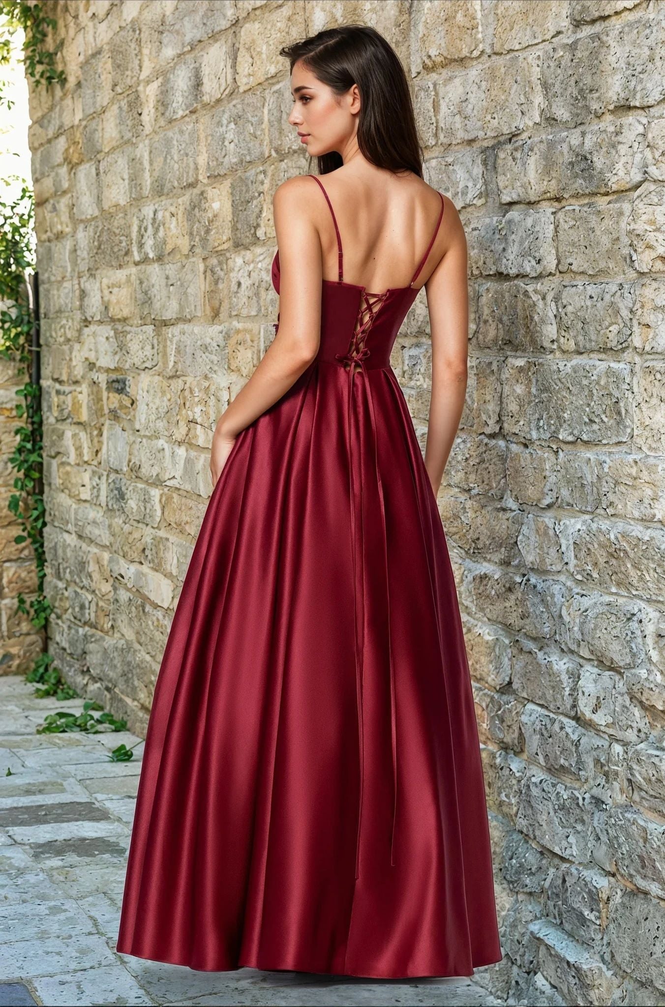 A woman stands outdoors by a stone wall in the Blondie Nites 5207BN, a sleeveless, floor-length burgundy satin prom dress with a corset bodice and lace-up back. This elegant A-line gown is available for under $200.