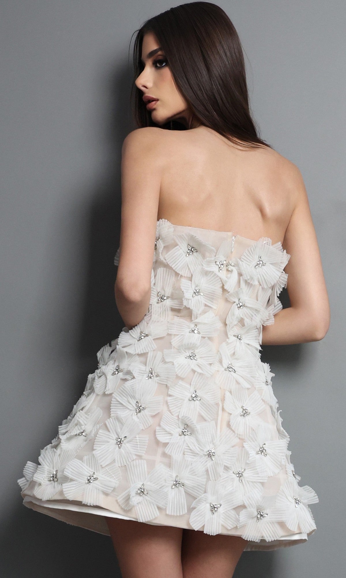 A woman stands facing sideways against a gray background, wearing the Jovani 52559 short homecoming dress—an elegant white strapless design embellished with large, textured 3D floral appliques.