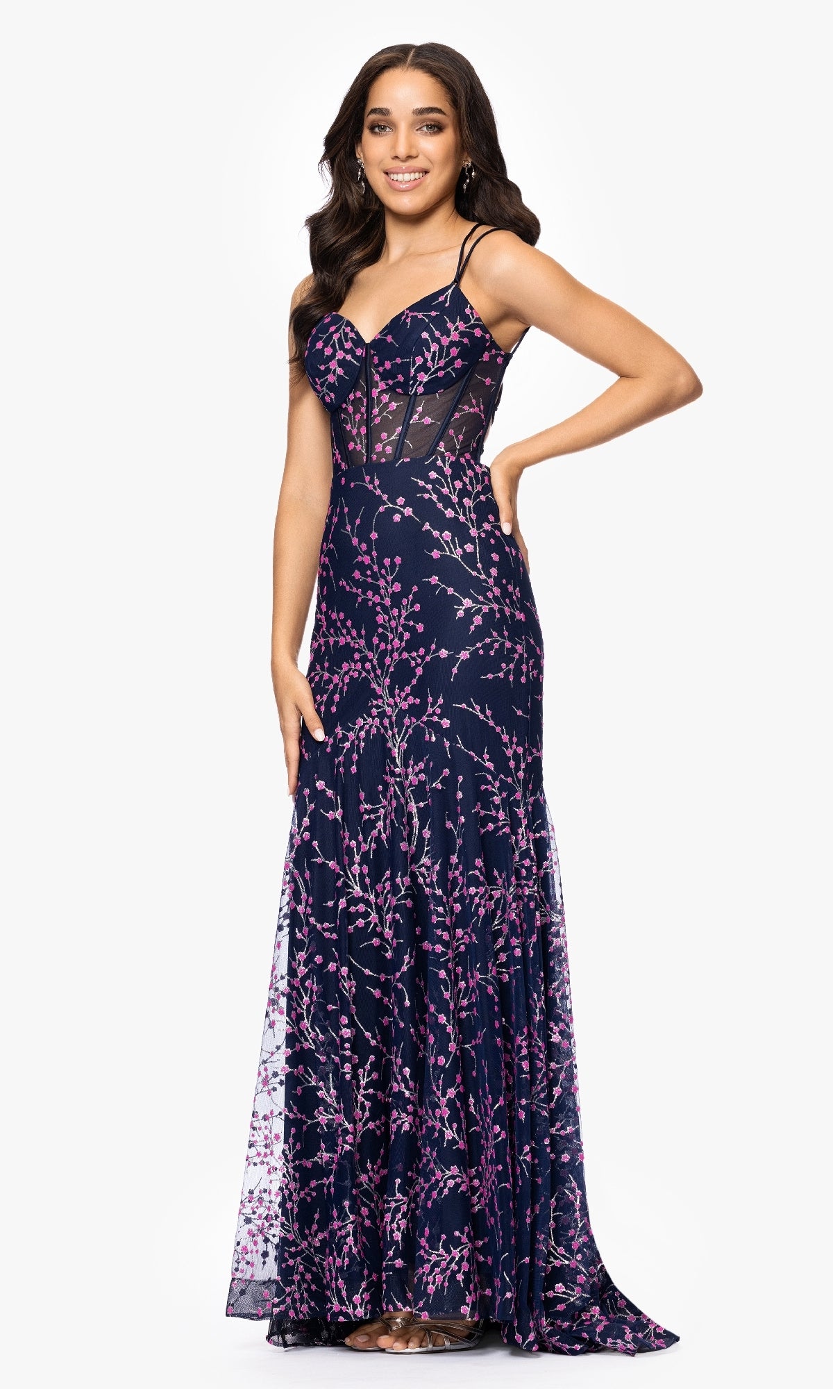 Woman wearing the Blondie Nites 5273BN Navy Blue Print Prom Dress, featuring thin straps, sheer panels, and pink floral patterns, stands and smiles with one hand on her hip.