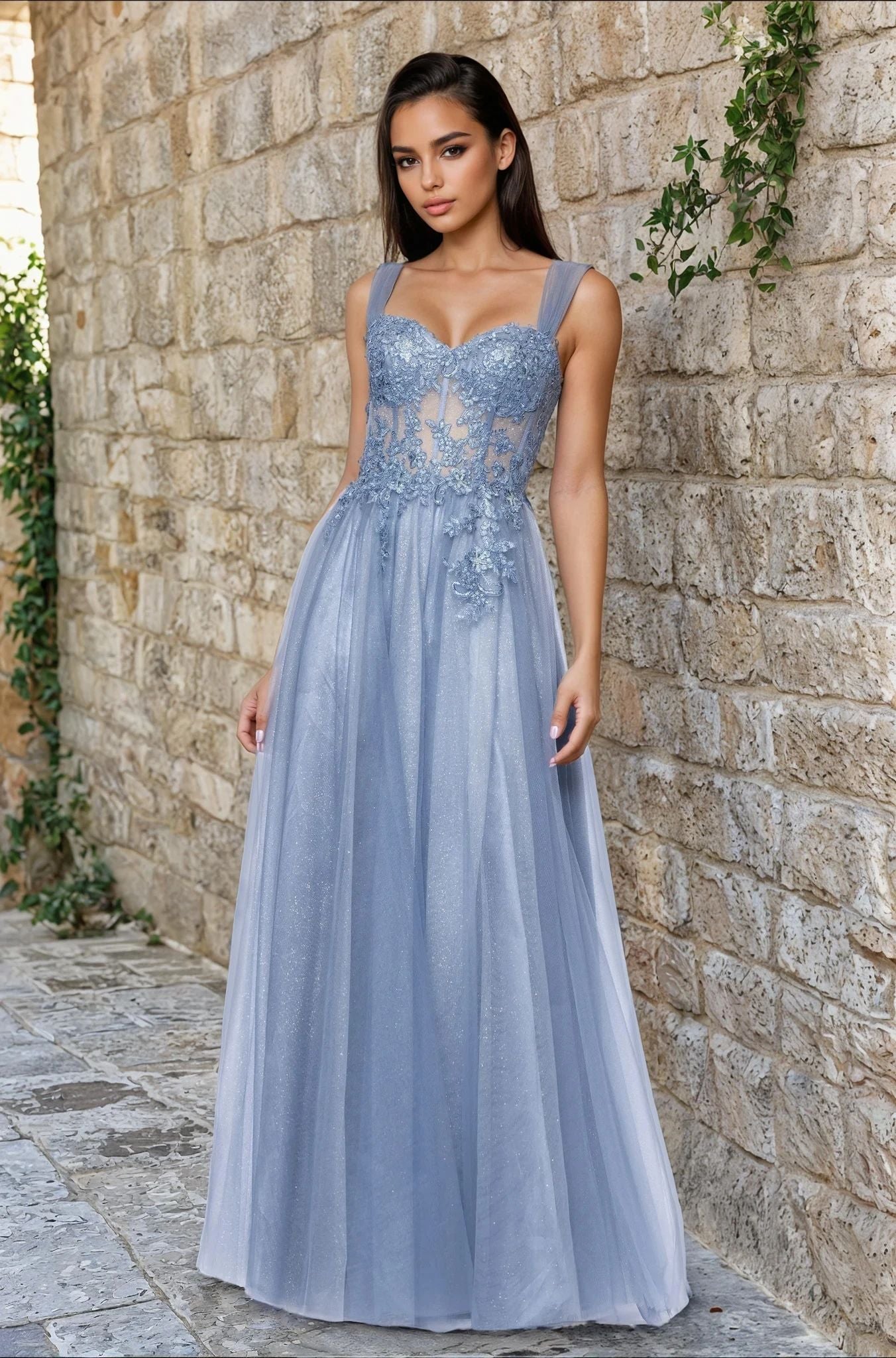 A woman in the Long Prom Dress: Blondie Nites 5311BN, a slate sequin-lace gown with a corset bodice, stands by a stone wall with greenery, gazing at the camera.