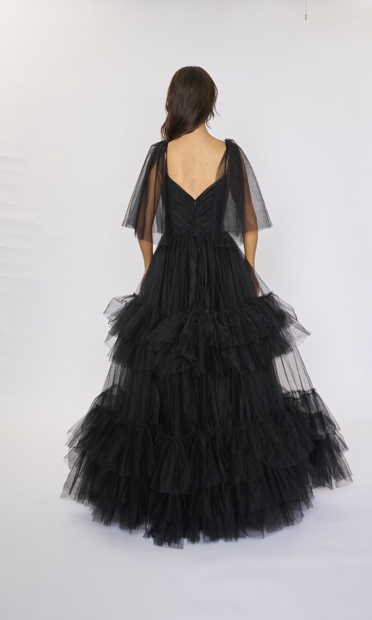 A woman is photographed from the back against a white background wearing the Nina Canacci 5315 long formal dress, featuring black ruffle-tiered tulle and short sheer sleeves.