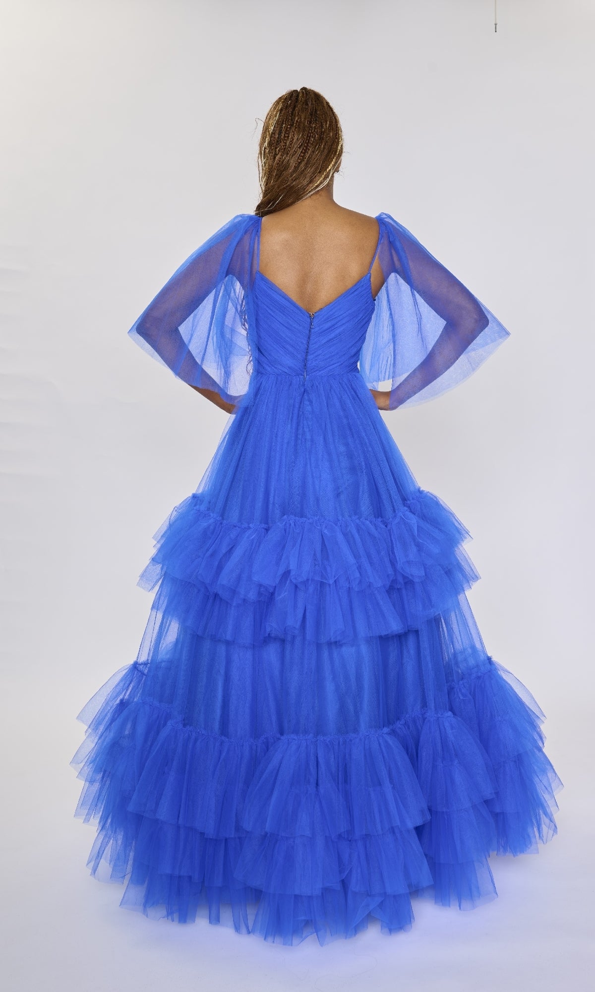 A woman faces away in a Nina Canacci 5315 bright blue long formal dress with ruffled tulle tiers and sheer, voluminous sleeves, set against a plain white background.