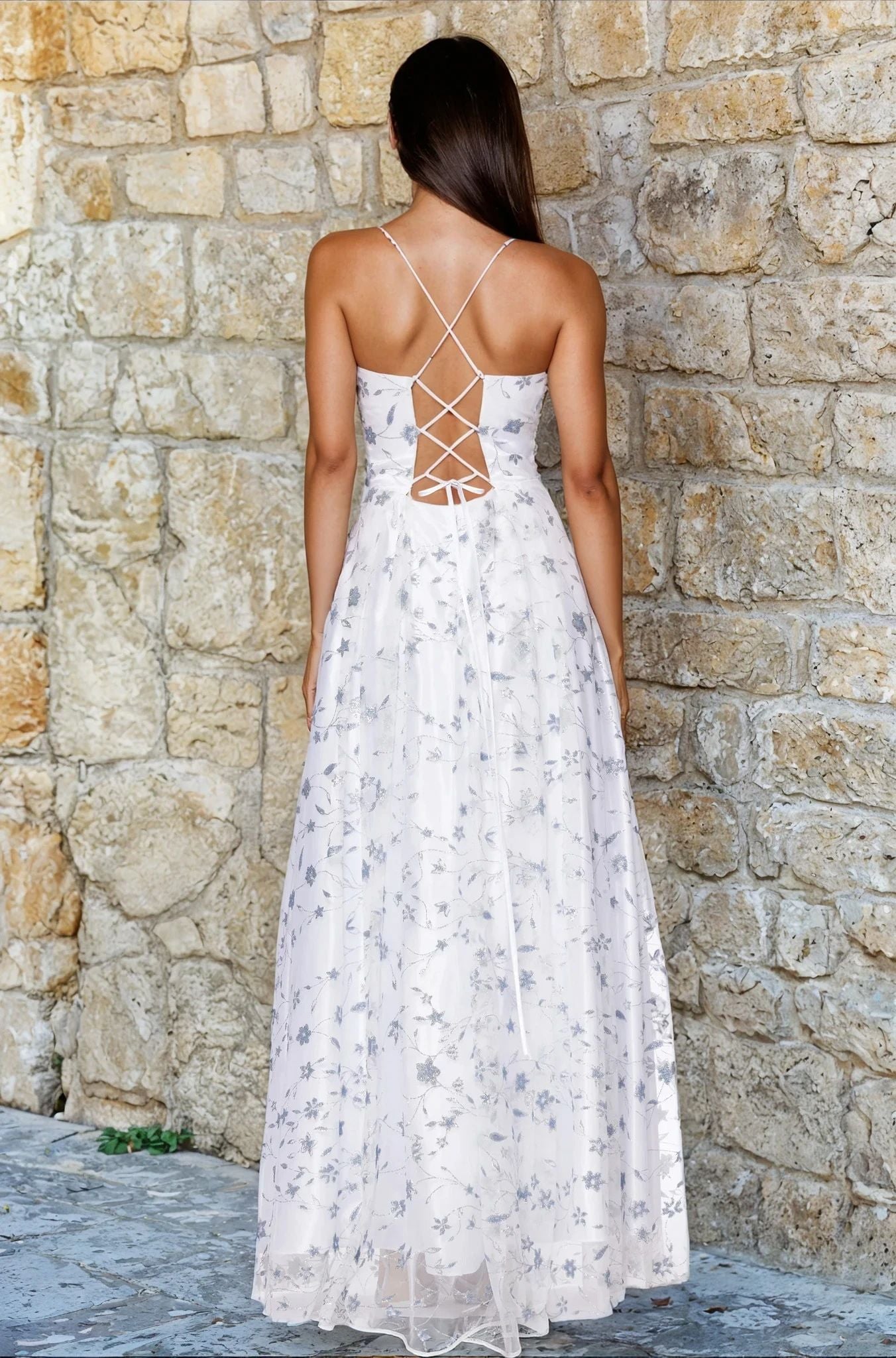 A woman wearing the Long Glitter Prom Dress: Blondie Nites 5317BN, featuring a white floral print and crisscross lace-up back, stands in front of a stone wall with her back to the camera.