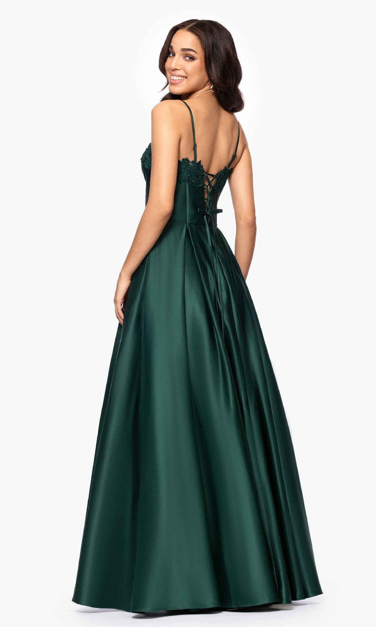 A woman stands against a plain white background, seen from the back, wearing the Classic Long Prom Dress: Blondie Nites 5387BN—a floor-length dark green satin gown with embroidered-lace corset details and thin straps.