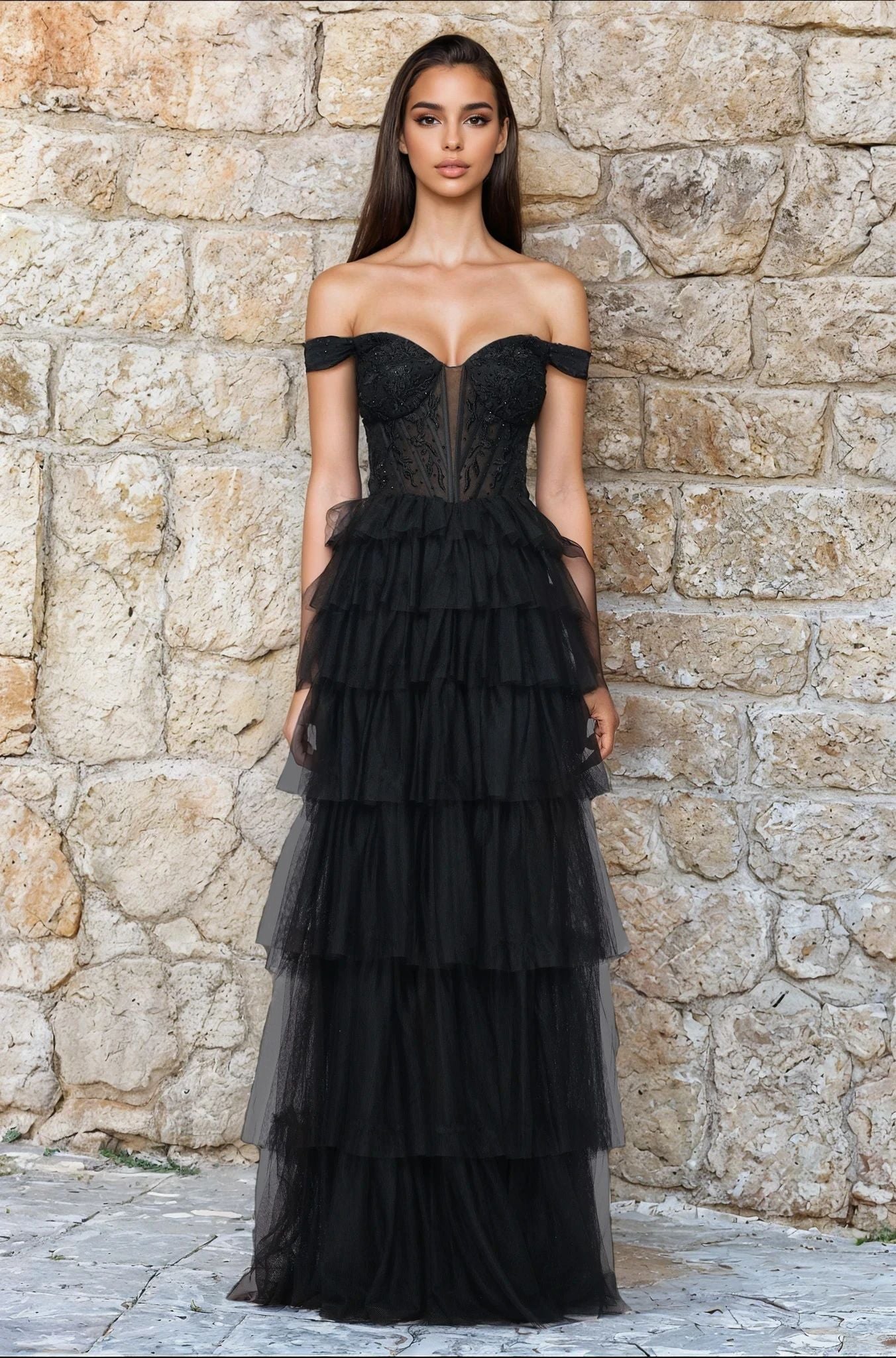 A woman stands facing forward, hands by her sides, wearing the Blondie Nites 5390BN Long Black Ruffle Prom Dress with off-the-shoulder tiered tulle ruffles, posed before a textured stone wall.
