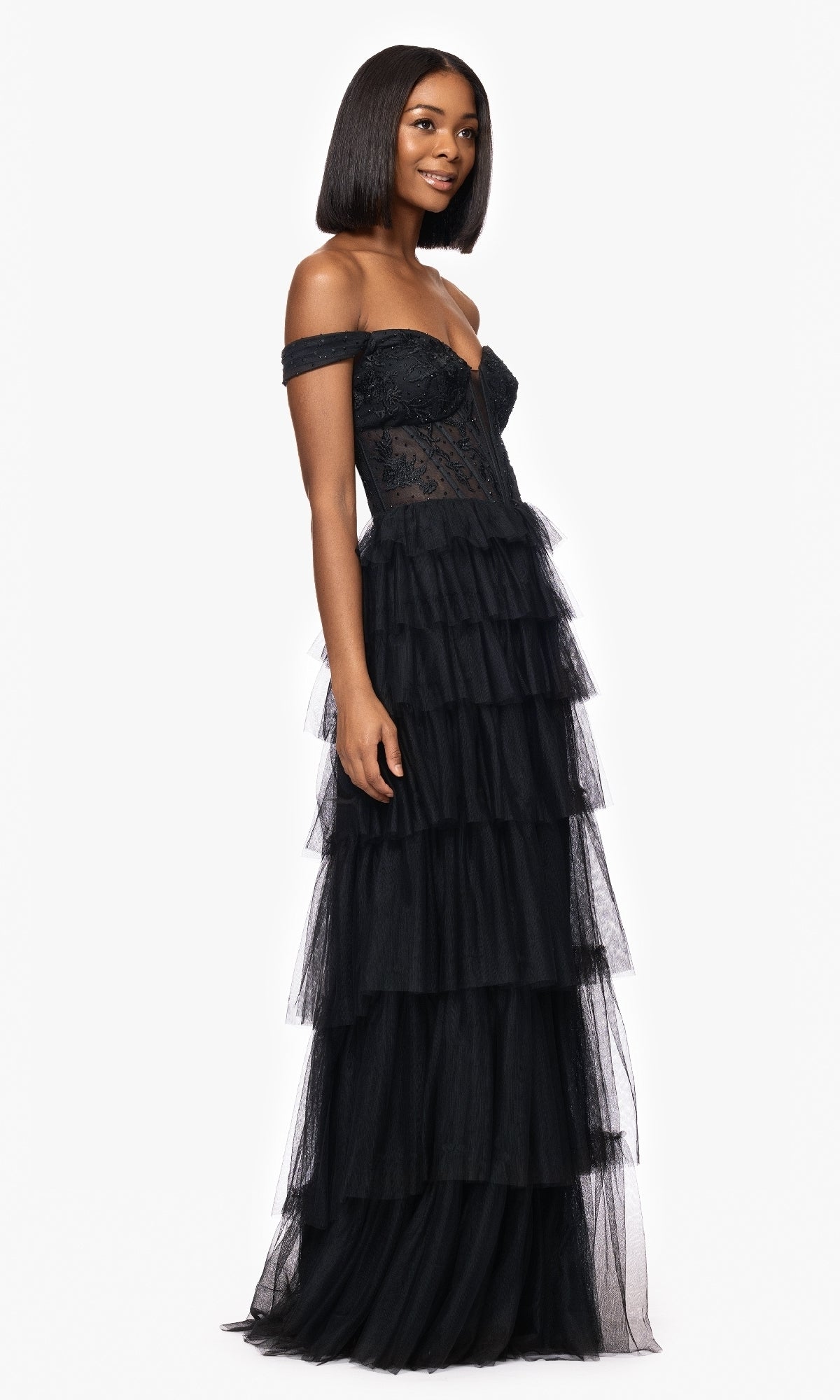 A woman stands against a white background wearing the Long Black Ruffle Prom Dress: Blondie Nites 5390BN, featuring an off-the-shoulder neckline, fitted bodice, and tiered tulle skirt.