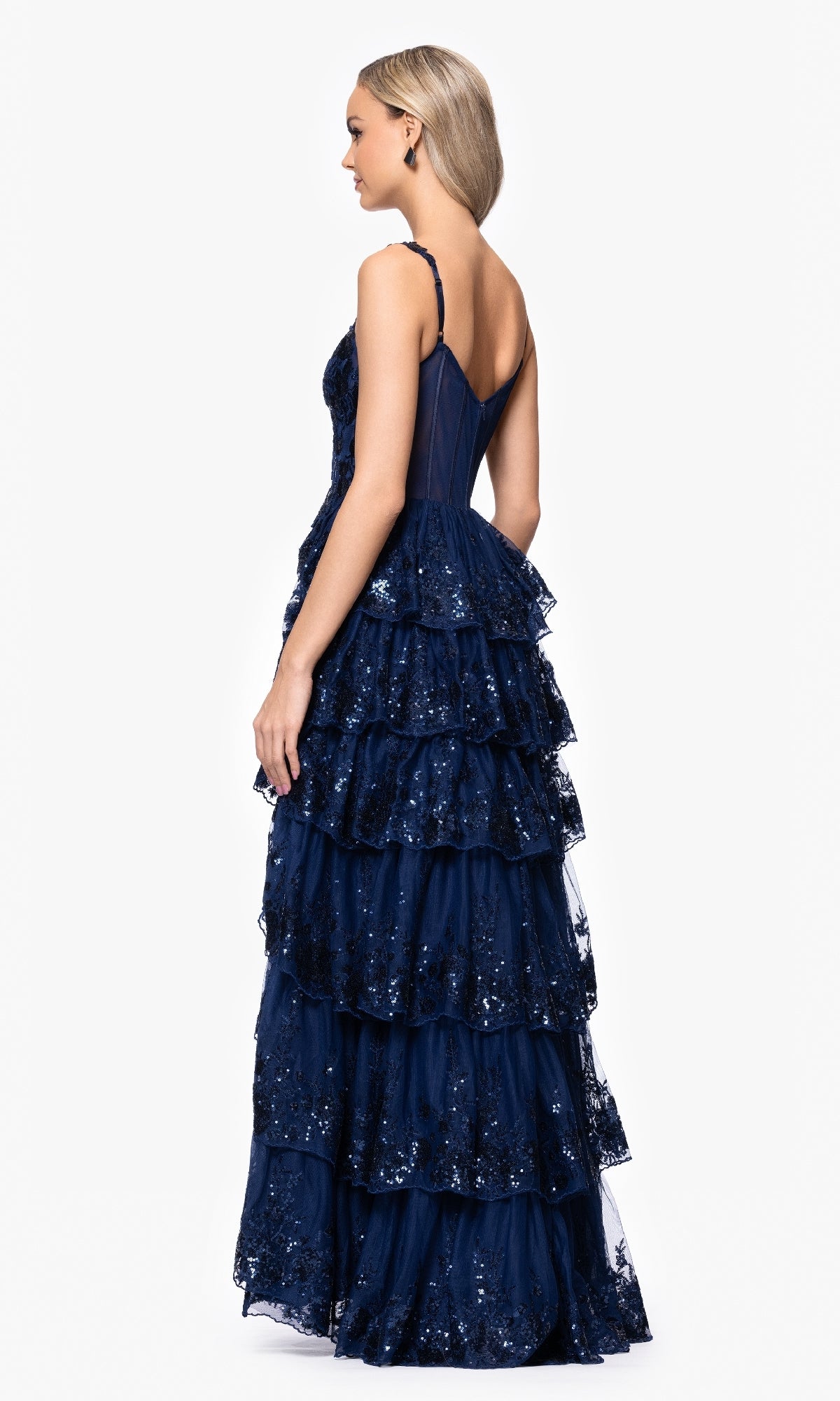 A woman wears the Long Navy Prom Dress: Blondie Nites 5394BN, featuring sleeveless sequin details and a tiered A-line skirt, standing slightly turned to showcase its elegant layered back design.