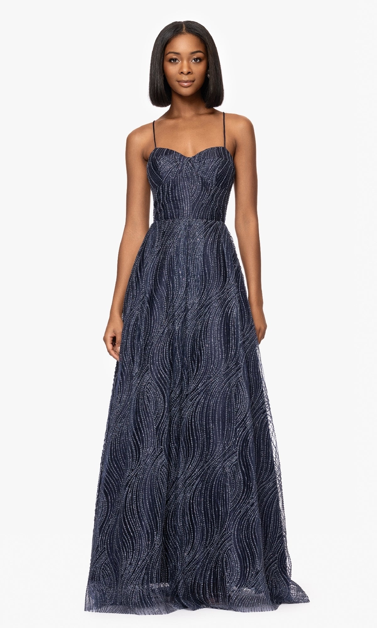 A woman wears the Long Navy Blue Prom Dress: Blondie Nites 5416BN, a floor-length gown with spaghetti straps and shimmery wavy patterned fabric, standing against a plain white background.
