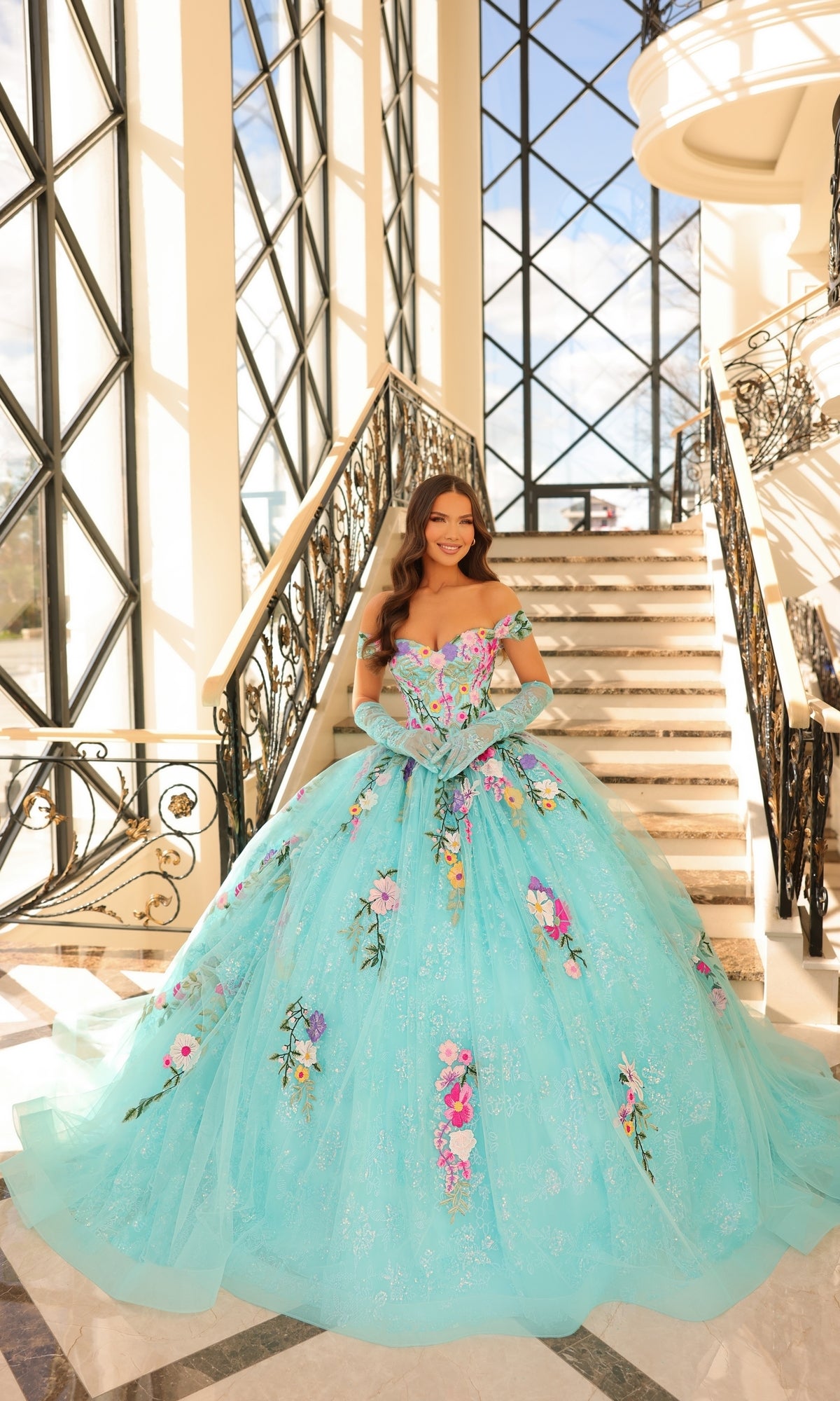 A woman wears the Quinceanera Dress 54200 By Amarra, a large light blue tulle ball gown with floral embroidery, as she stands on marble stairs in a sunlit, elegant interior with tall windows.