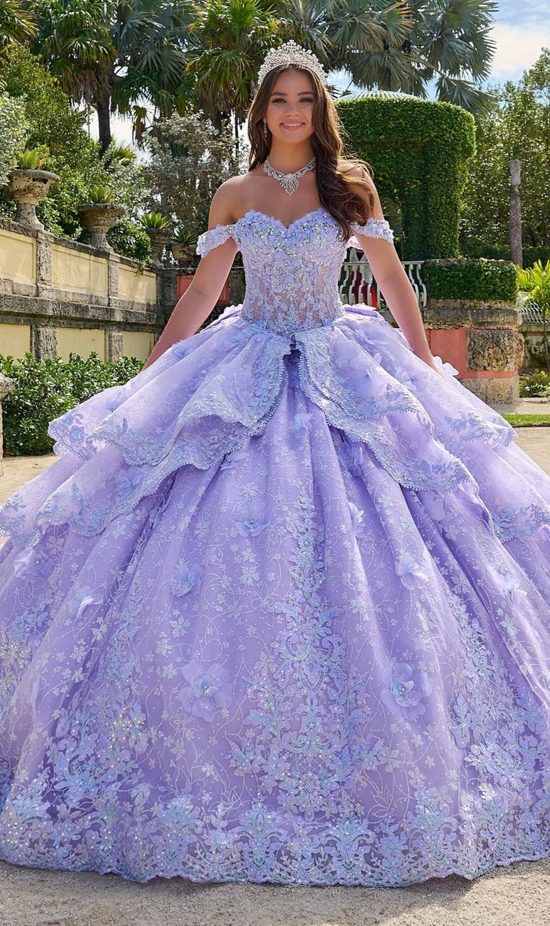 A young woman stands outdoors in a garden wearing the Quinceanera Dress 54202 by Amarra, an off-the-shoulder lavender ball gown with lace embroidered flowers, paired with a tiara and jeweled necklace.