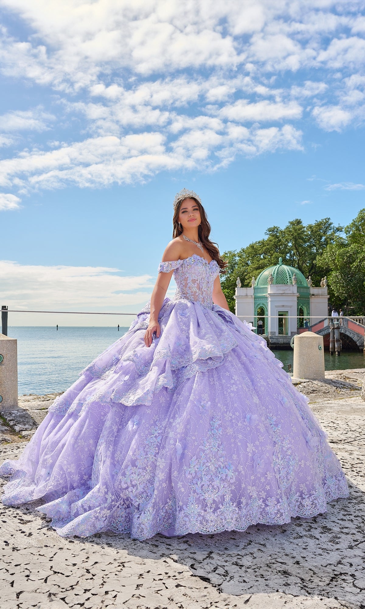 A young woman stands outdoors near a waterfront, wearing the Quinceanera Dress 54202 By Amarra—a lavender ball gown with off-the-shoulder sleeves and lace embroidered flowers—with trees and a domed building in the background.