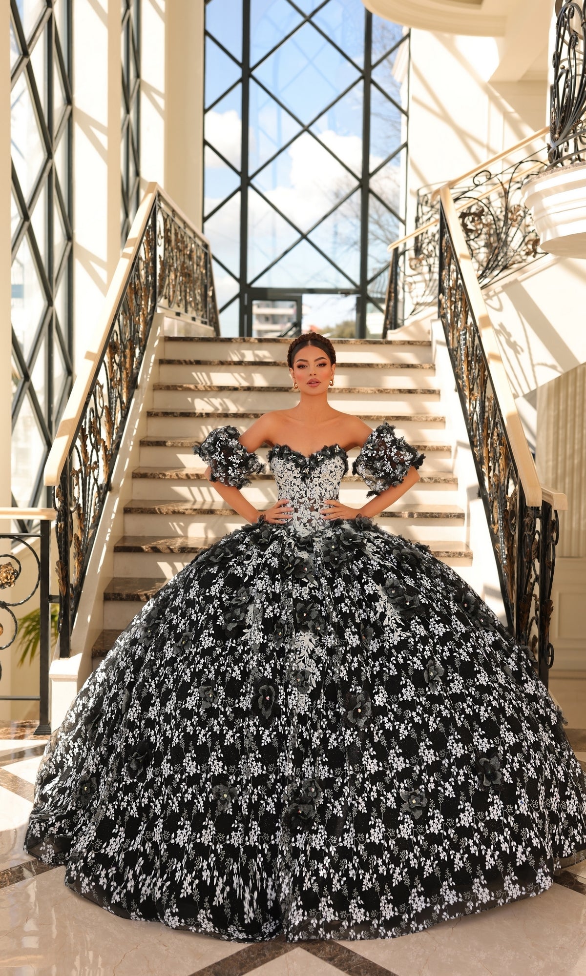 A woman in the Quinceanera Dress 54206 by Amarra, featuring glow-in-the-dark floral lace and a strapless sweetheart neckline, stands on a marble staircase in a sunlit, elegant interior with large windows and ornate railings.