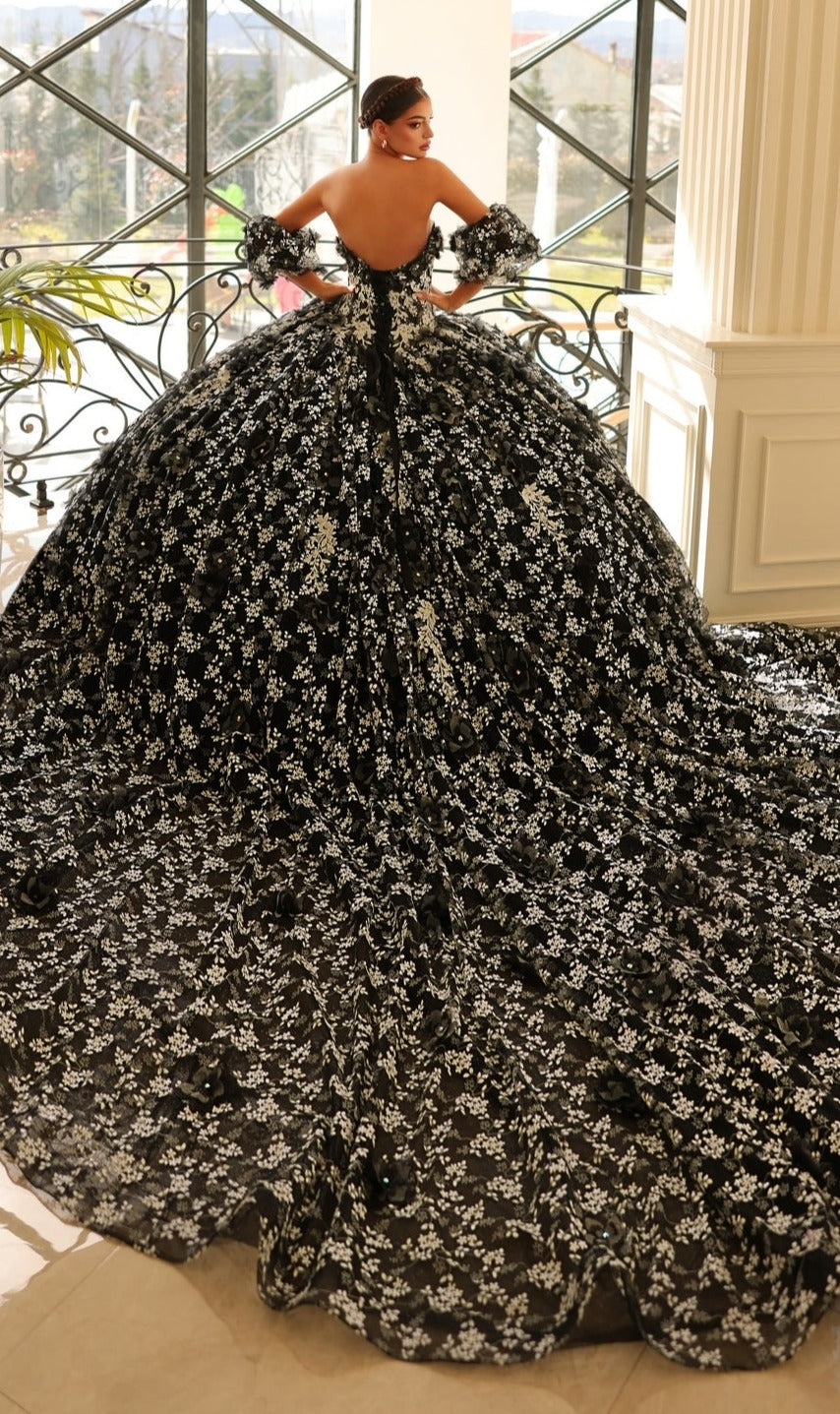 Indoors by a window, a person wears the Quinceanera Dress 54206 By Amarra—a dramatic off-the-shoulder ball gown with black and white floral patterns and a sweeping train.
