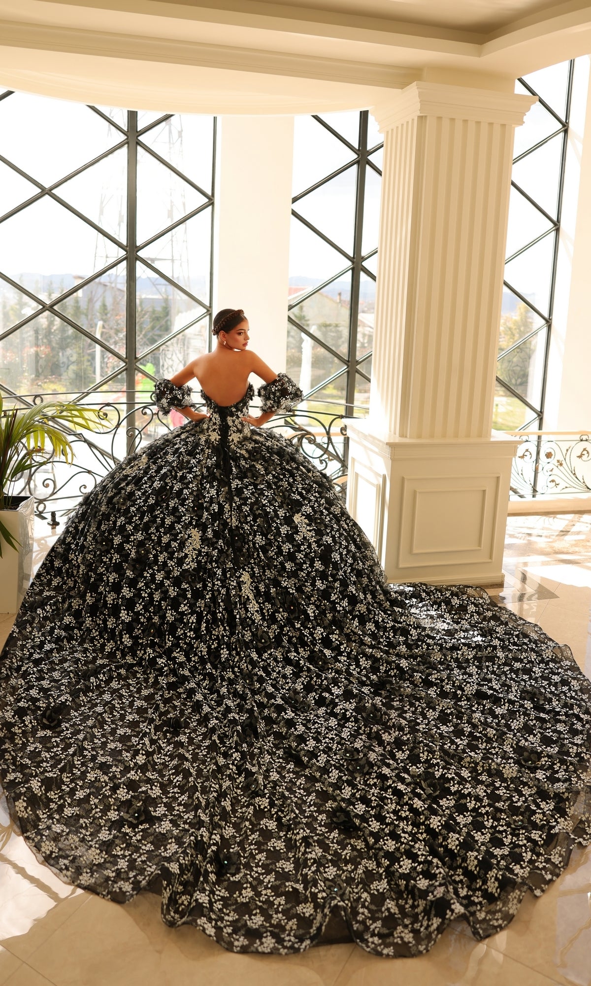 A woman wearing the Quinceanera Dress 54206 By Amarra, featuring white glow-in-the-dark floral lace, stands indoors near tall windows and a column.