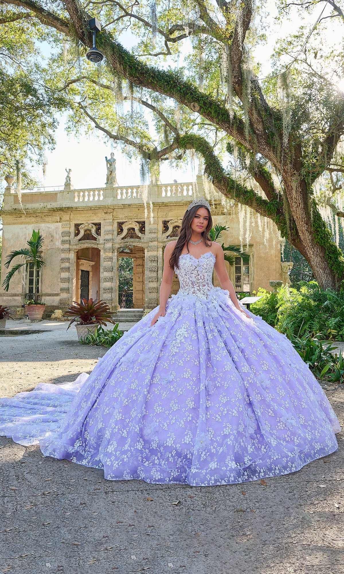 A woman stands outdoors in front of a historic building, wearing the Quinceanera Dress 54206 by Amarra—a stunning lavender ball gown with white floral details and a long train.