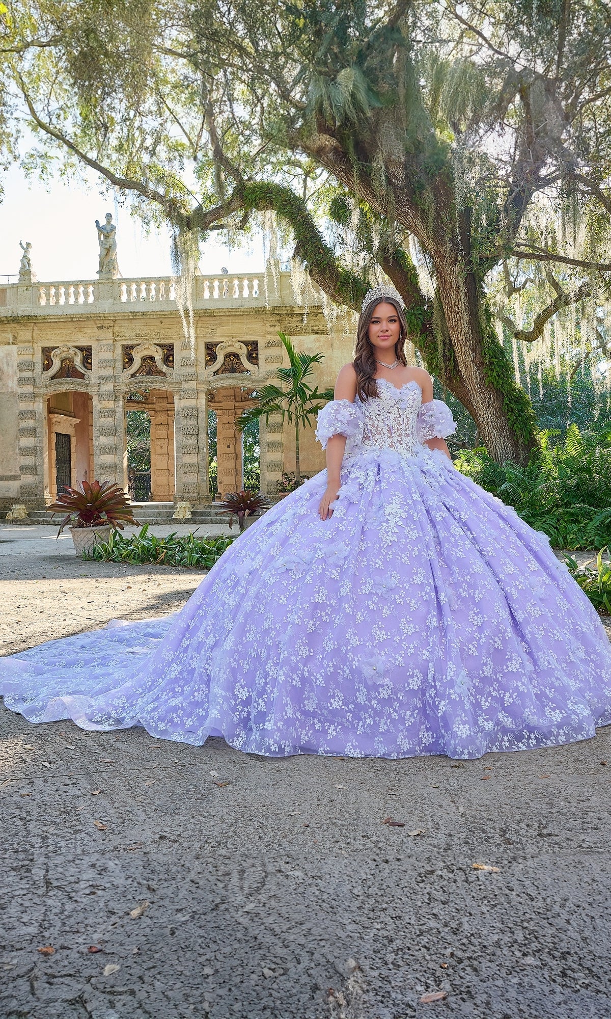 A young woman stands outdoors before a historic stone building, wearing the Quinceanera Dress 54206 By Amarra—a stunning lavender ball gown adorned with white floral details.