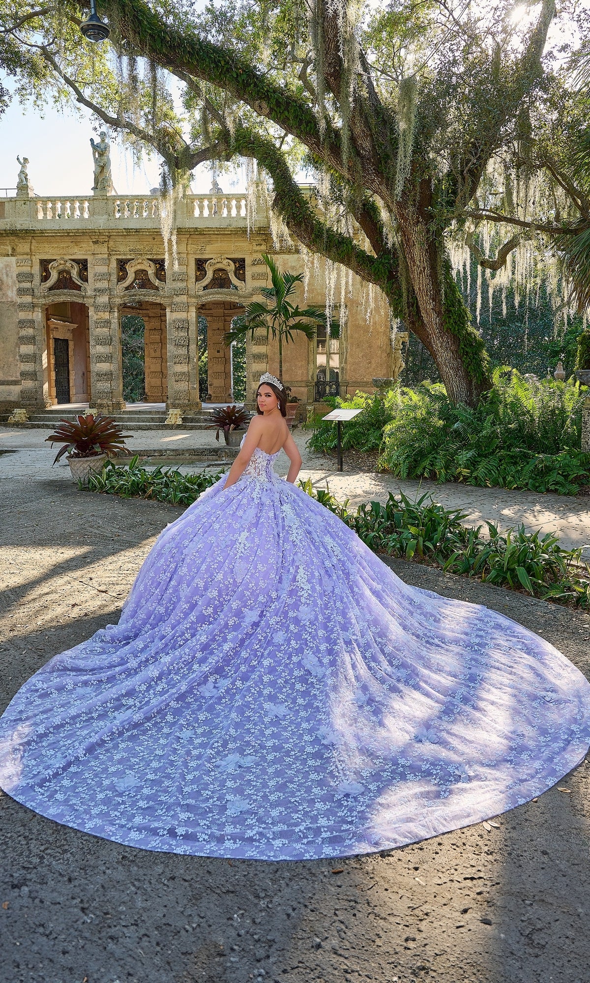 A woman wearing the Quinceanera Dress 54206 By Amarra stands on an outdoor path, her long train flowing behind her with a historic building in the background.
