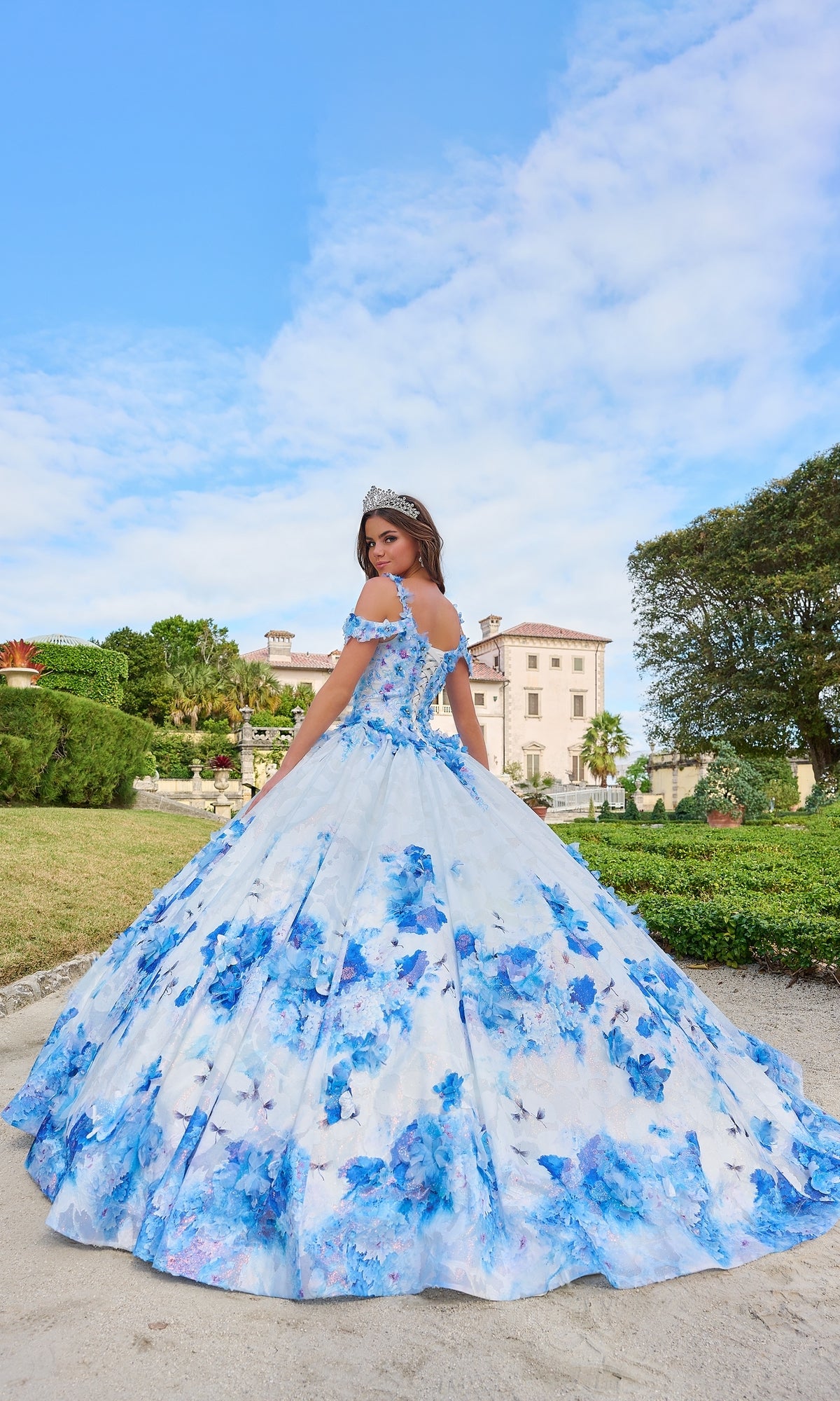 A woman in the Quinceanera Dress 54208 By Amarra, a blue and white floral ball gown with a tiara, stands outdoors before a grand mansion and trees under a partly cloudy sky.