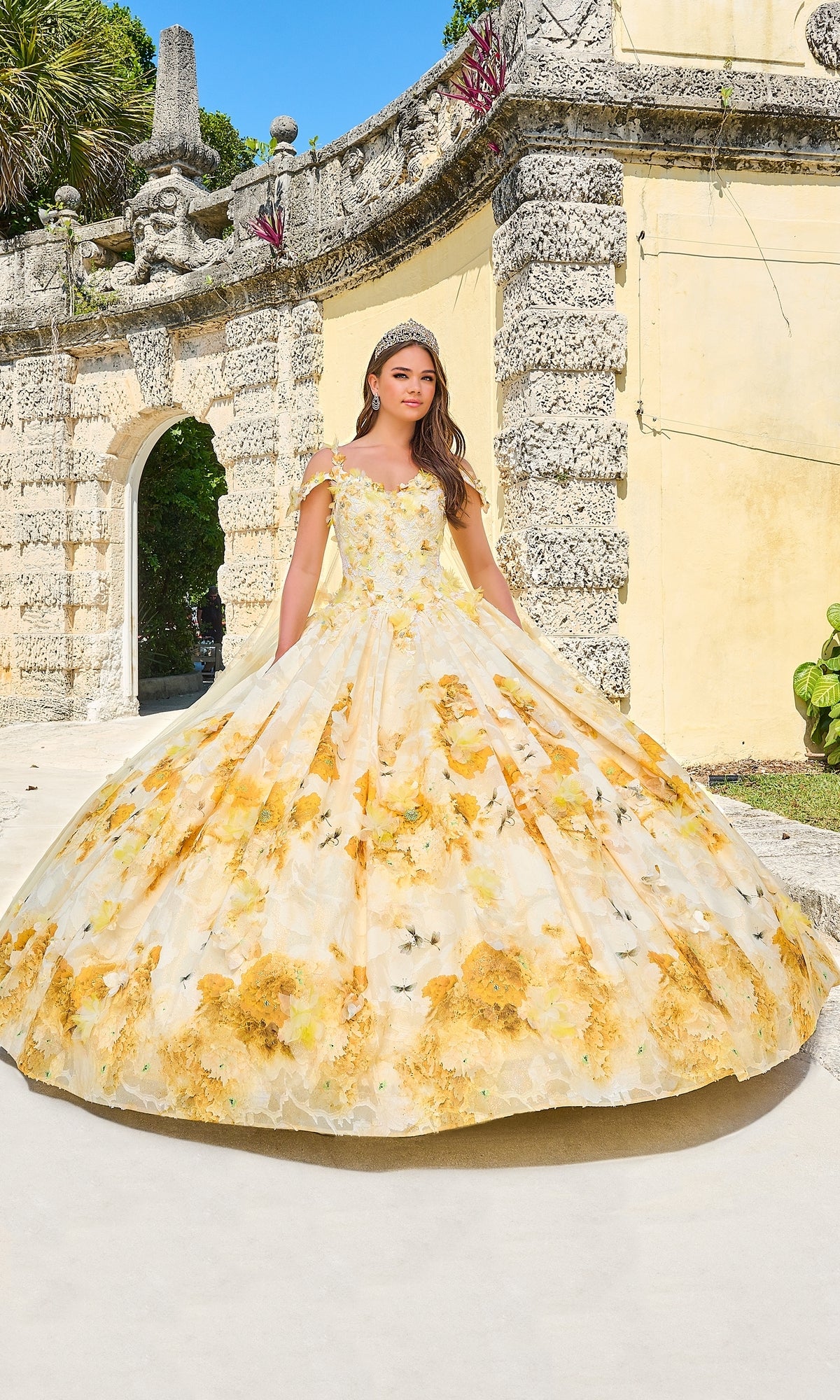 A young woman poses outdoors in the Quinceanera Dress 54208 By Amarra, a glitter tulle ball gown with floral print and an off-the-shoulder design, set against a stone wall and lush greenery.