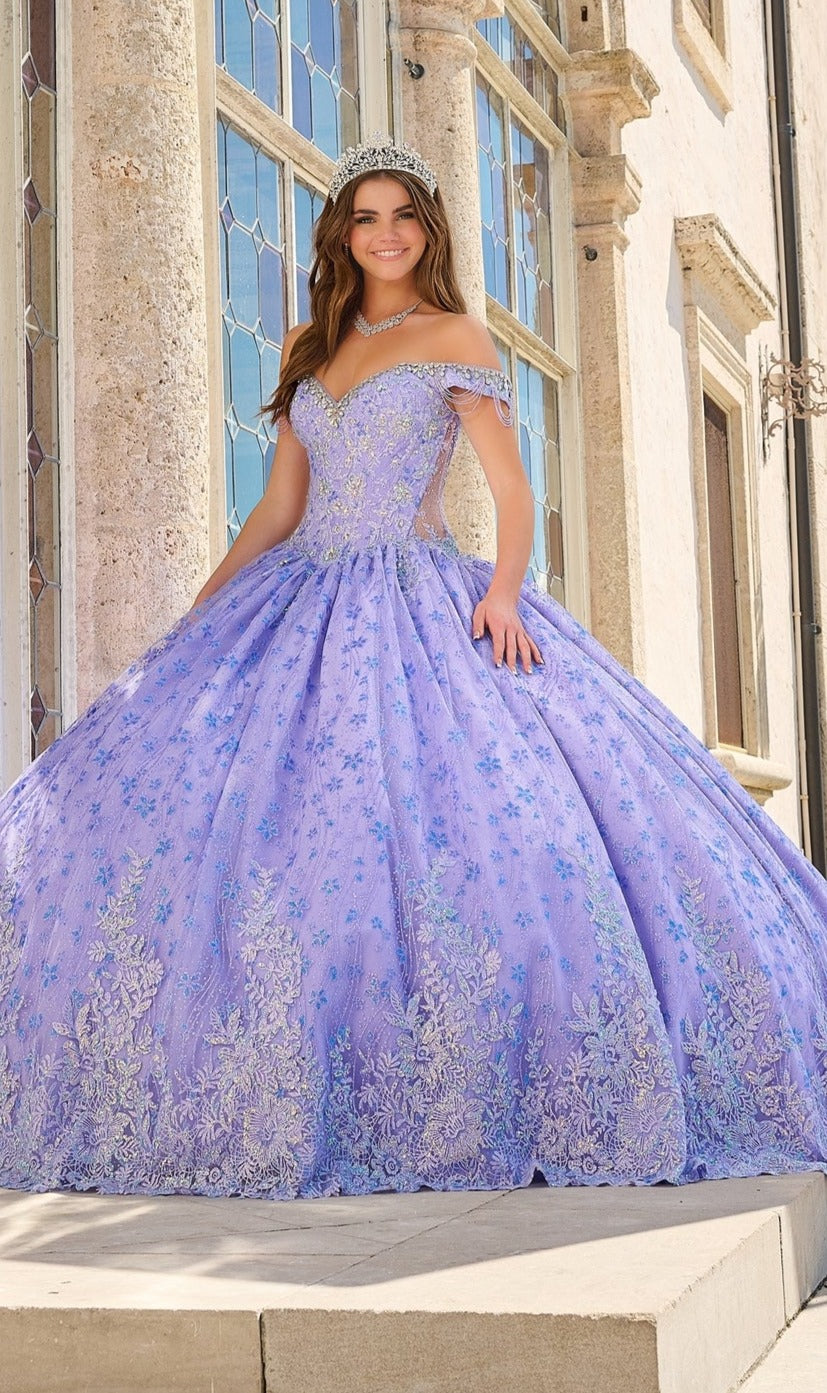 A woman in the Quinceanera Dress 54220 by Amarra, a lavender off-the-shoulder ball gown with floral glitter tulle embroidery and a tiara, stands before a stone building with large windows.