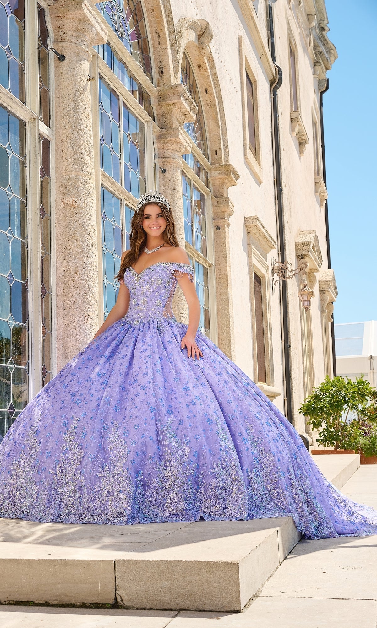 A young woman wears the Quinceanera Dress 54220 by Amarra, an off-the-shoulder lavender ball gown with floral glitter tulle, while standing outside a stone building with arched windows on a sunny day.
